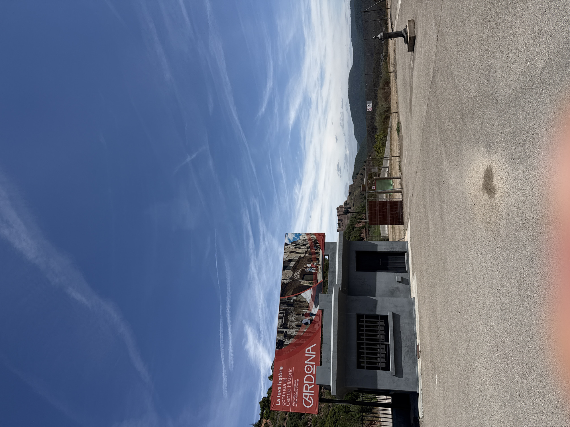 The media file captures an outdoor scene with a clear blue sky featuring wispy white clouds. In the foreground, a modern grey building with dark-trimmed windows stands on a paved area. A large, red banner with white lettering is prominently displayed on the building's facade. The banner reads "La teva història continua al Centre Històric" and "Your story continues in the Historic Centre," with "CARDONA" in large white letters. Below this, there's a smaller line of text in English. The banner also features images of historical architecture. To the left of the building, a chain-link fence encloses some greenery. Further back, a gentle slope leads to hills covered in trees and vegetation, with some structures visible on the hillside. A dark, ornate water fountain with a square base is situated on the paved area to the left of the building. The overall impression is a bright, sunny day, likely in the afternoon given the sunlight and shadows, suggesting a historical site or tourist attraction in Cardona, Spain.