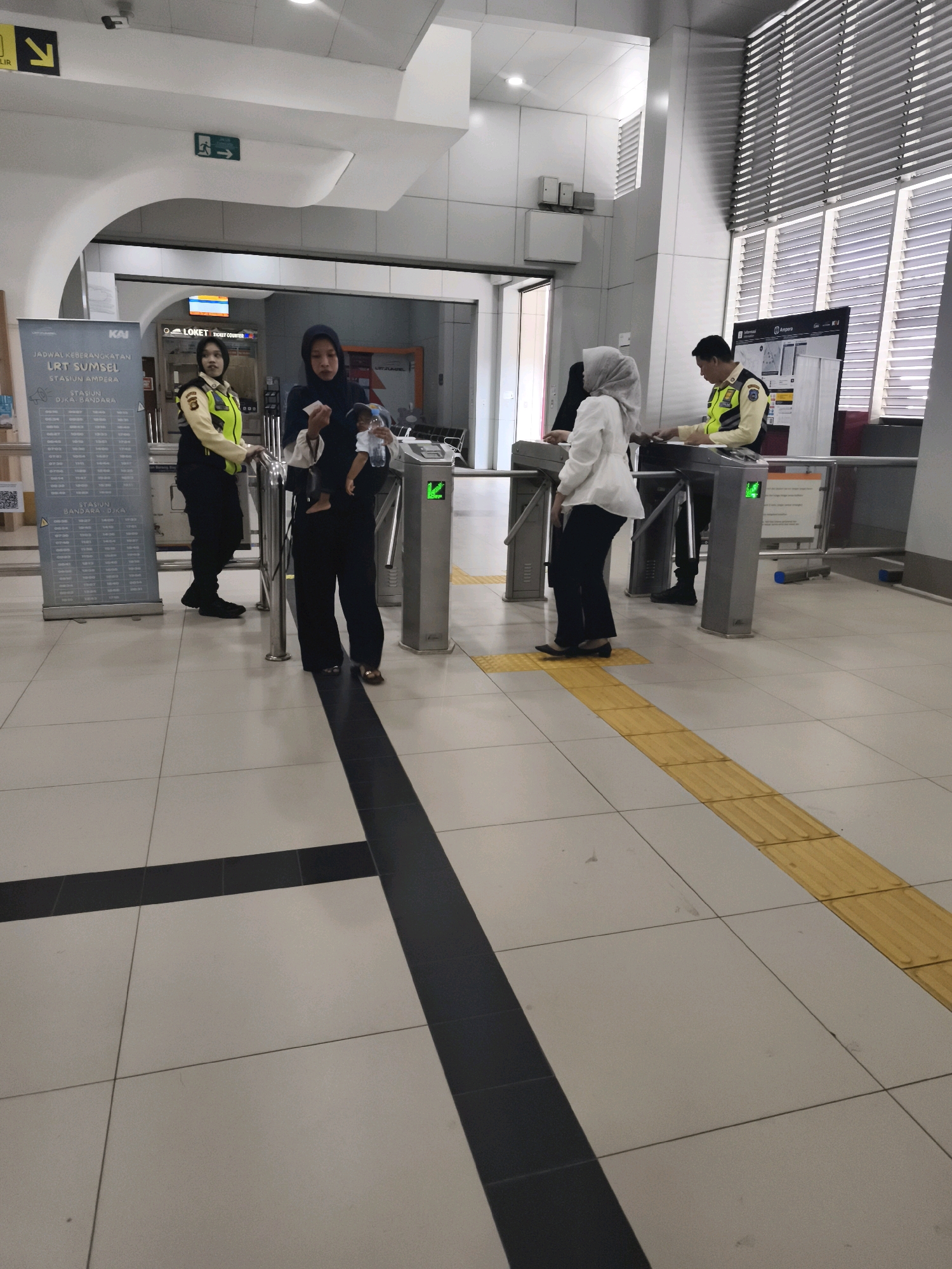 This image depicts the interior of a modern transportation hub, likely a train or light rail station, in the vicinity of Kelurahan 16 Ilir, Kec. Ilir Timur I, Indonesia. The scene is characterized by a tiled floor with distinct black and yellow accent lines, suggesting a clear pathway. Several individuals are present, including two uniformed security personnel, one on the left wearing a yellow vest over a dark uniform and another on the right in a similar vest. Two civilians are interacting with turnstiles; one woman, wearing a dark hijab and clothing, is holding a young child and a water bottle, appearing to pass through a turnstile. Another woman, in a white hijab and outfit, is also near a turnstile. In the background, there are signs, ticket counters, and architectural elements of the building, including a patterned ceiling and ventilation grilles. The lighting appears to be artificial.