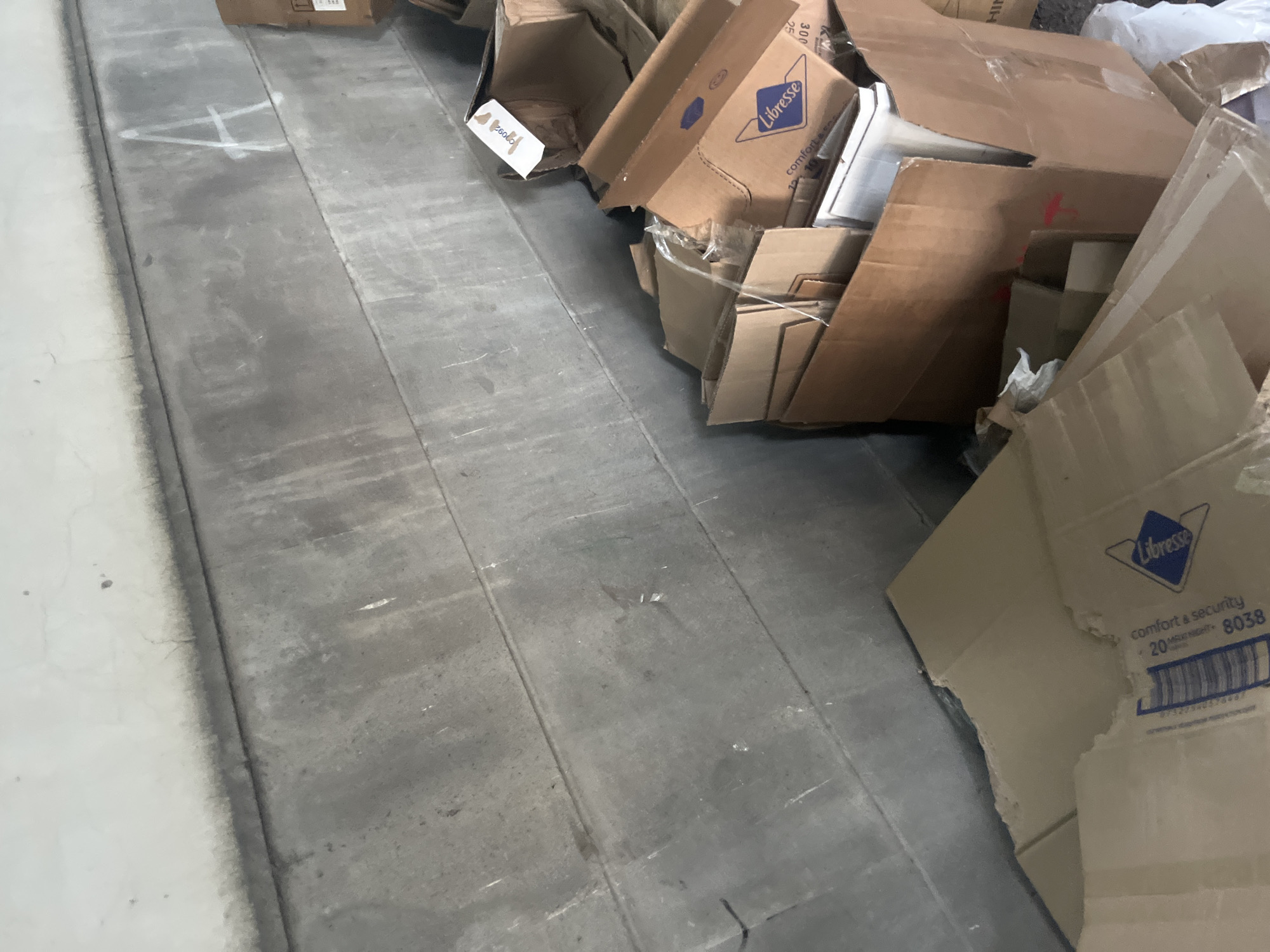 The image shows a collection of flattened cardboard boxes, some bearing the brand name "Libresse," scattered on a gray concrete floor. The location is not specified, and there is no indication that the image was taken in Yerevan, Armenia. There are no people visible, and the focus is solely on the discarded cardboard. The environment is a simple interior space with artificial lighting implied by the evenness of illumination.