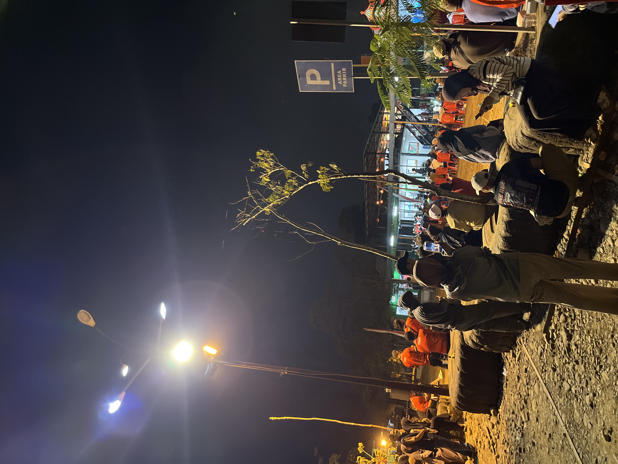In the city of Unknown, Indonesia, a group of people are gathered outdoors at night. The scene is illuminated by streetlights and lights from a building in the background. The people are dressed in casual clothing, and some are sitting on large rocks or tires. Others are standing and observing the surroundings. In the background, a building with an open-air seating area is visible, with many people seated at tables. A parking sign is also present in the frame. The ground is covered with gravel and rocks. The overall atmosphere suggests a social gathering or event taking place in an informal outdoor setting.