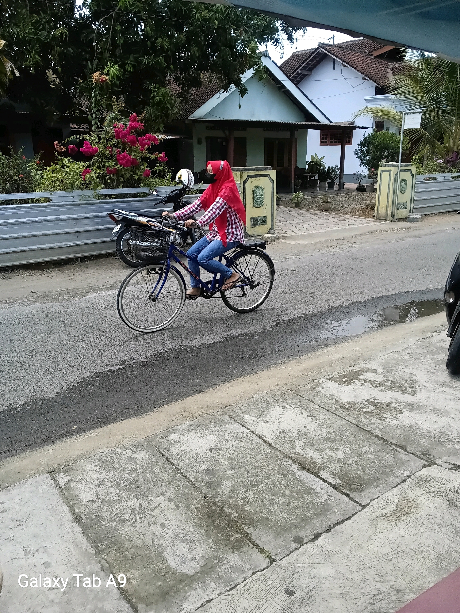 A person wearing a red hijab and a checkered long-sleeved shirt rides a blue bicycle down a paved road in the city of Karangsoko, Indonesia. The person is also wearing blue jeans and appears to have a dark mask on their face. The bicycle has a black basket on the front and the wheels are visible, along with the person's feet on the pedals. A parked motorcycle is visible behind the bicycle. The background shows a white house with a tiled roof, trees, and a fence. The road appears wet in places, suggesting recent rain. There is also a concrete sidewalk on the right side of the road. The image also displays a watermark at the bottom left that reads "Galaxy Tab A9".