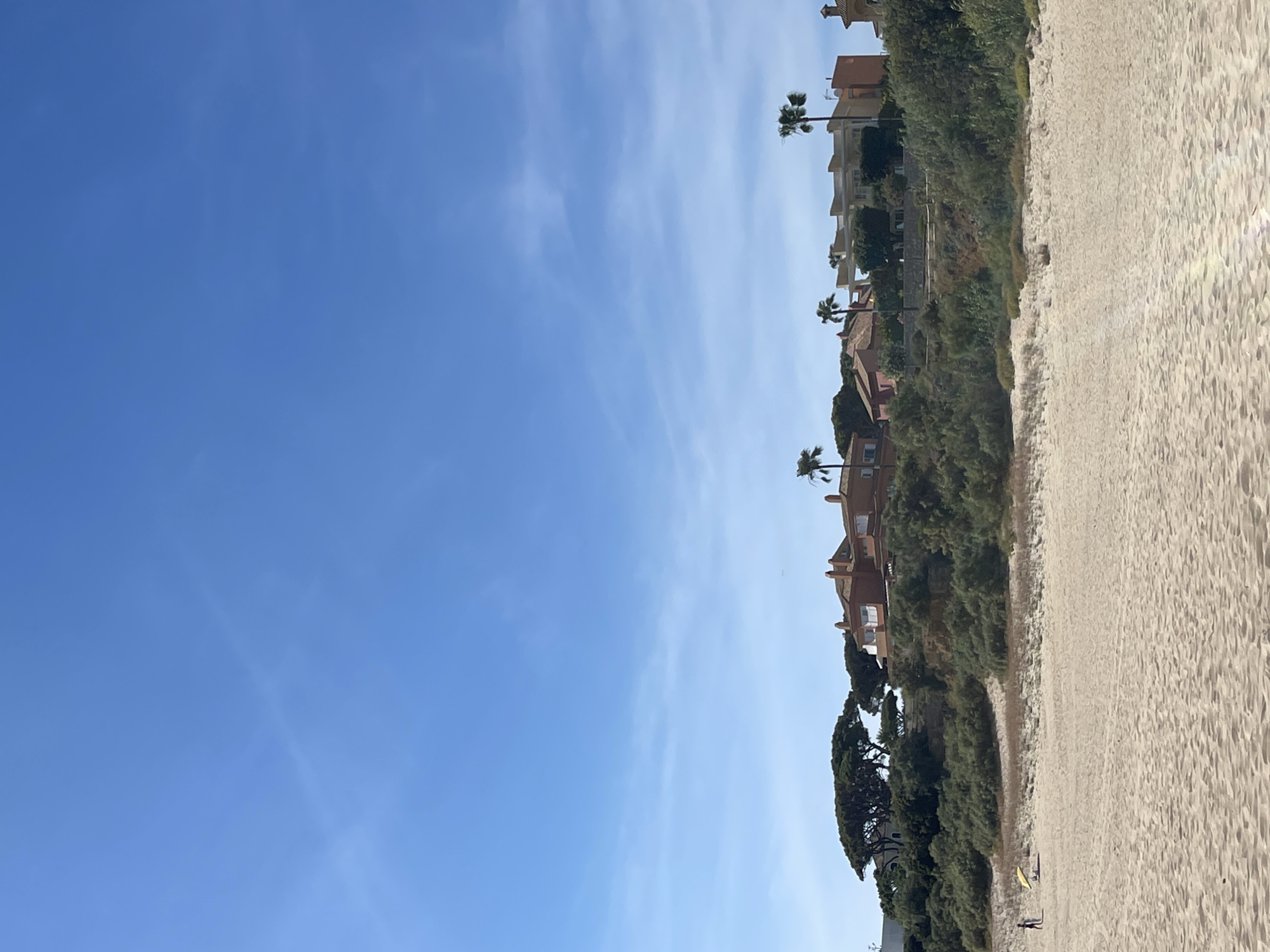 A sideways outdoor shot shows a sandy beach adjacent to a cliff with houses and palm trees. The beach occupies the right side of the image, with the cliff covered in trees and bushes separating it from the houses, which are primarily light brown with terracotta roofs. Several palm trees are interspersed among the houses. In the lower right corner of the image, two people are visible walking on the sand near the vegetation. A yellow object, possibly a surfboard, is next to them. The sky is a clear blue with some wispy clouds. Based on the architecture and vegetation, the location is likely the city of El Puerto de Santa María in Spain.