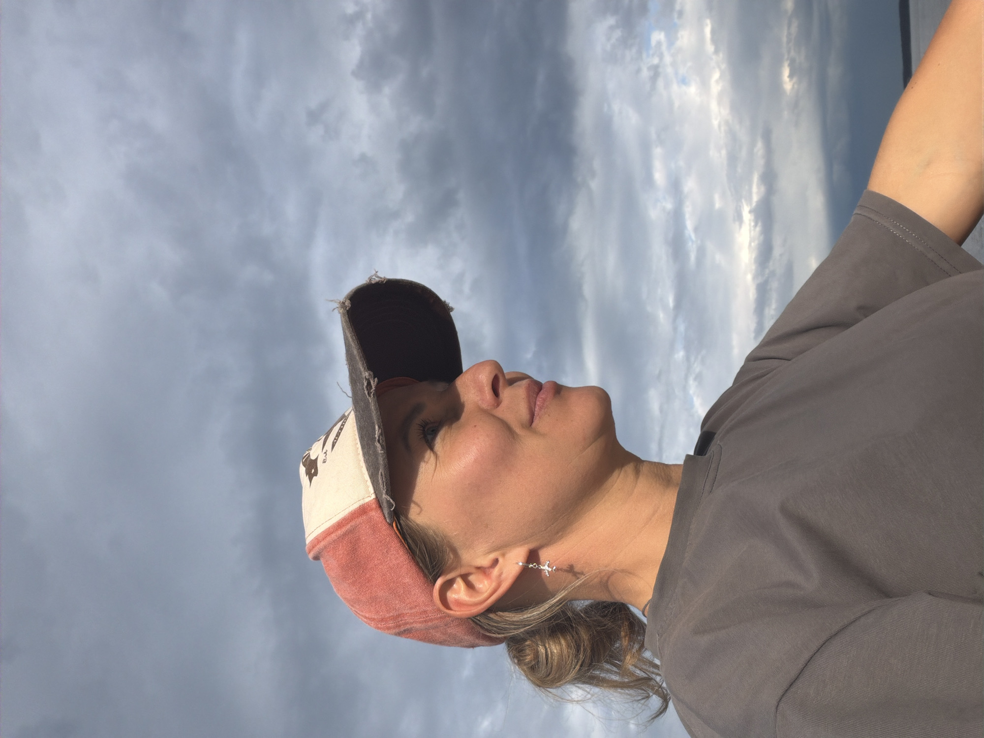 A medium shot shows a woman from the chest up, in profile, looking to the right, outdoors under a cloudy sky. She is wearing a grey t-shirt and a baseball cap. The cap is red with a beige front panel that has a dark graphic on it. Her hair is pulled back into a ponytail and she is wearing a silver earring shaped like a cross in her left ear. The background is dominated by a dramatic sky with dark, heavy clouds. The lower right of the image shows a sliver of water and a distant shoreline, suggesting the location is near a body of water. It is not possible to determine the specific location within Chaikovsky, Russia from this image alone.