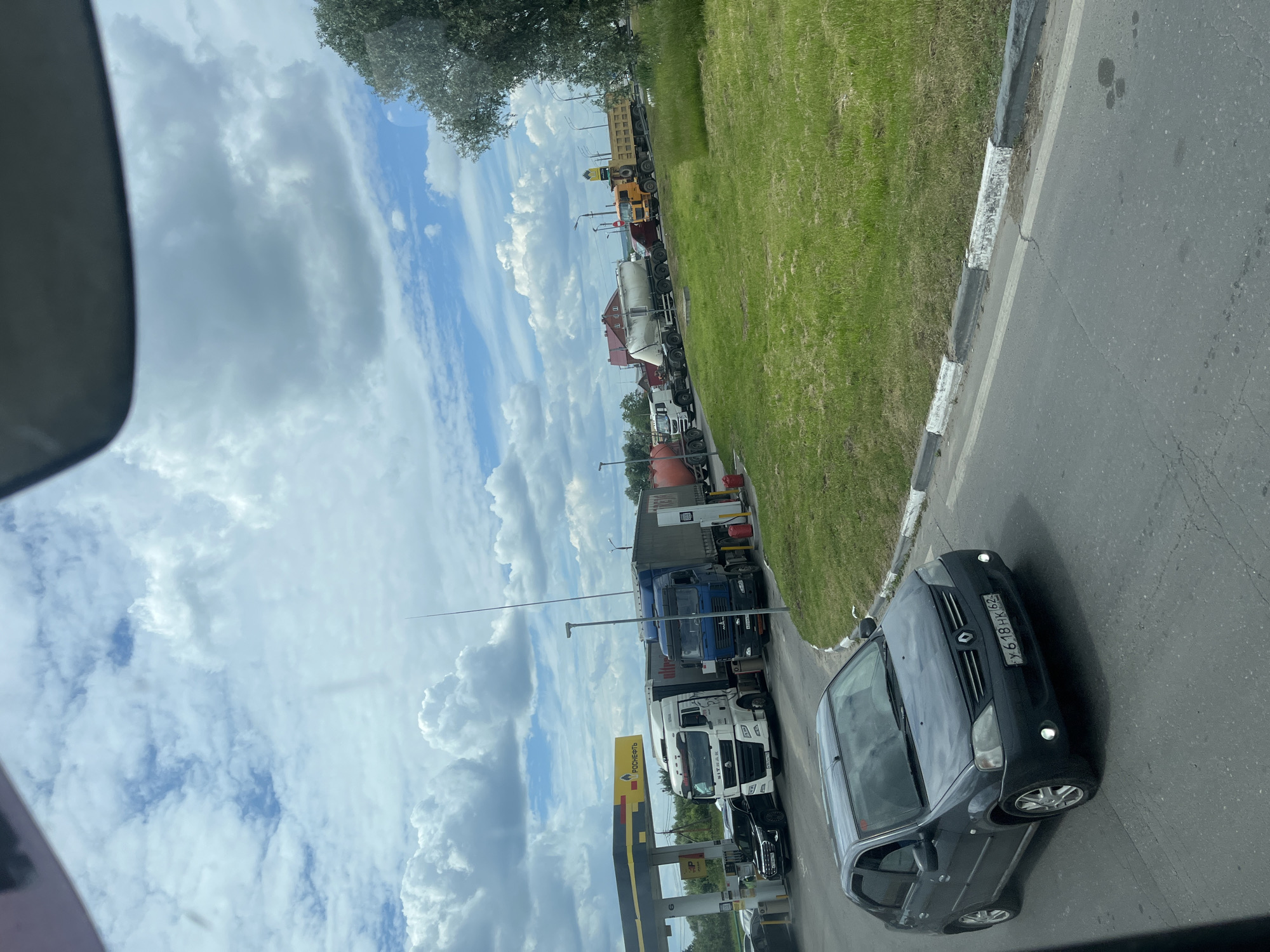 The image shows a queue of trucks and a car on a paved road next to a grassy area. The scene appears to be a roadside, possibly near a gas station, given the presence of a yellow and black sign that says "РАСПРОДАЖА". The sky is filled with white, fluffy clouds against a blue backdrop, suggesting daytime with fair weather. The trucks are of various types, including what appear to be cement mixers and flatbed trucks. In the foreground, a dark-colored car is visible with a license plate that reads "У 618 НК 162". The orientation of the image is rotated, making it appear as if the road is vertical and the sky is to the left. The overall impression is of a normal day with traffic on a road in what is suggested to be Старое Бобренево, Russia.