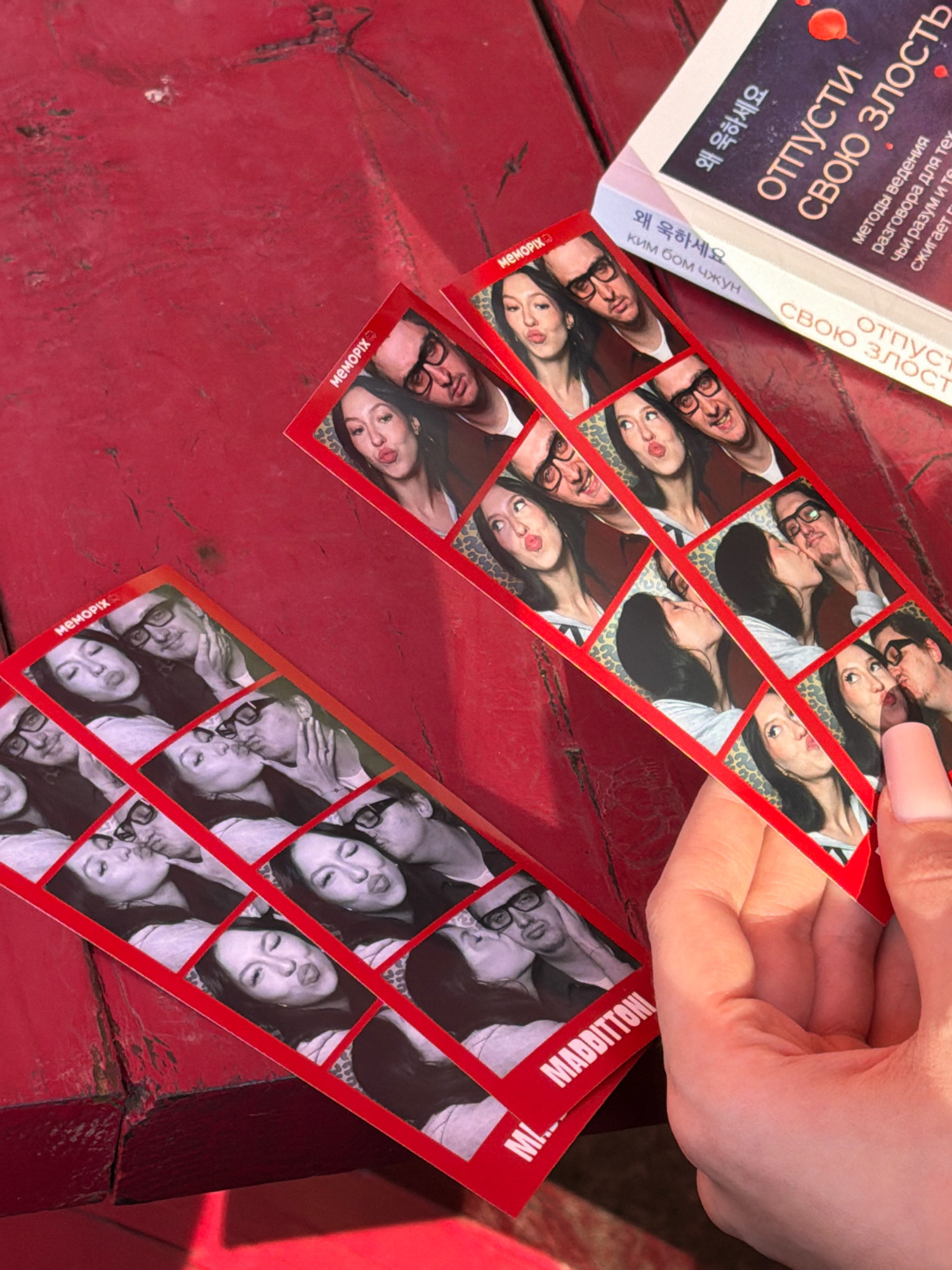 The image shows a close-up, overhead view of a red wooden surface, likely a table, with several photo strips and a book. The photo strips feature black and white images of a couple posing playfully. The book is open and displays text in Russian. The overall scene appears to be informal and personal. The specific location in Moscow, Russia is not determinable from the image.