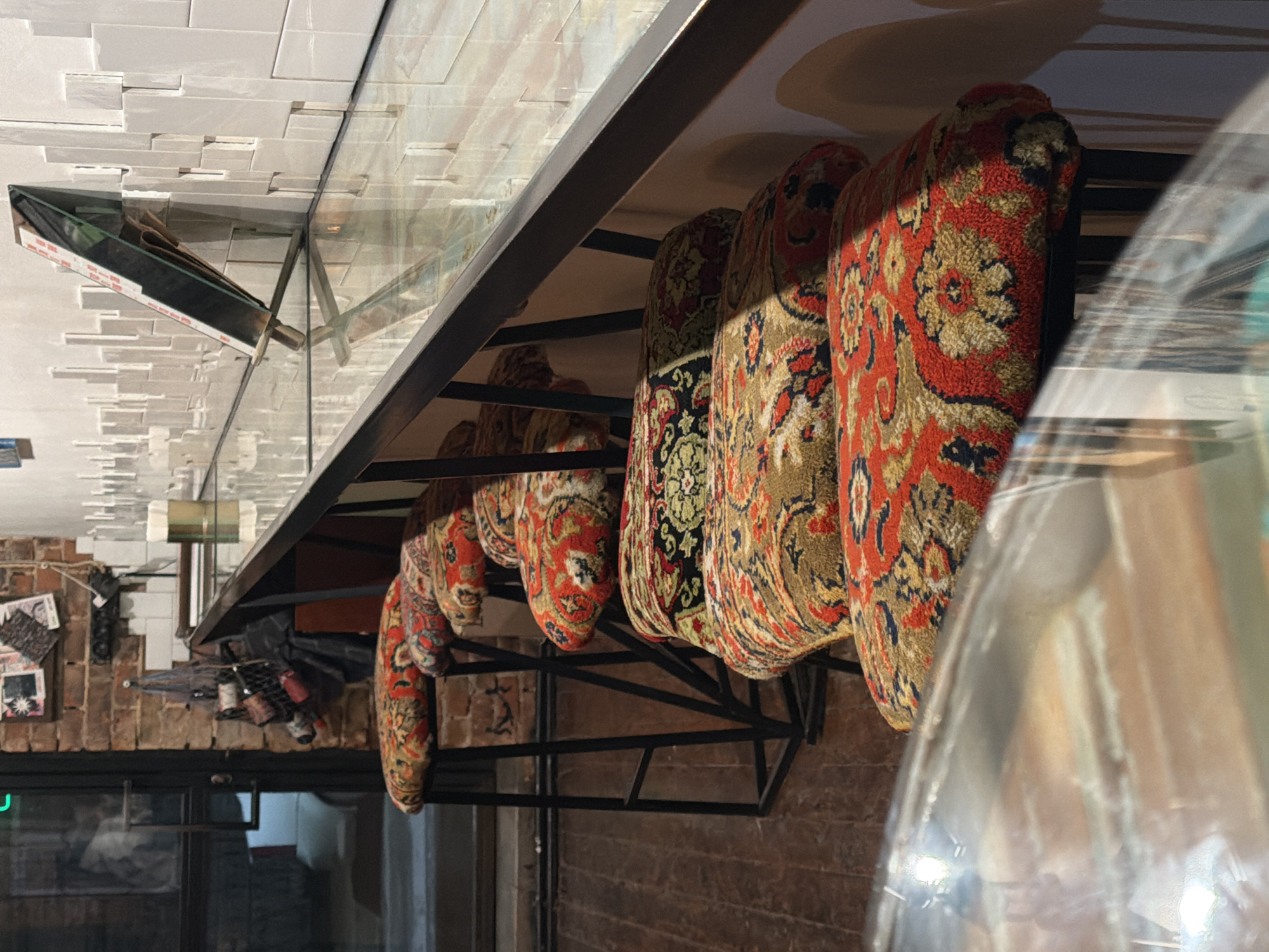 The image shows a shelf with several patterned cushions, likely in Rostov-on-Don, Russia, given the prompt. The cushions are suspended from a black metal rack attached to a brick wall, with a glass surface above the cushions reflecting the ceiling and other objects. The environment includes white brickwork and tiles in the reflection, along with other objects like bottles and framed pictures. The lighting is subdued, and the composition includes some reflections and distortions.