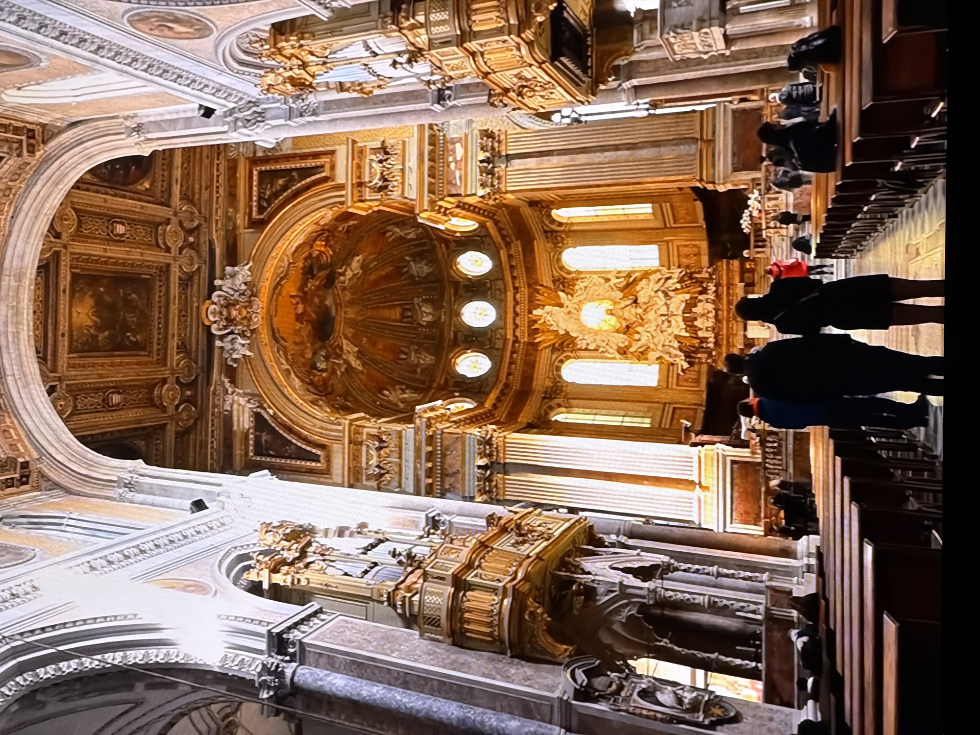 The image depicts the interior of a grand cathedral. The architecture is ornate and features high ceilings with intricate golden ornamentation and painted scenes. Large columns line the nave, and gilded balconies or pulpits are visible on either side. The altar area is prominent, with a highly detailed sculpture or altarpiece illuminated by natural light from windows. Several pews are arranged in rows, with some people seated and others standing. In the foreground, a few individuals are visible from behind, facing towards the altar. The scene conveys a sense of solemnity and grandeur typical of a religious establishment. The image does not show any elements that indicate the location is Chirchik, Uzbekistan.