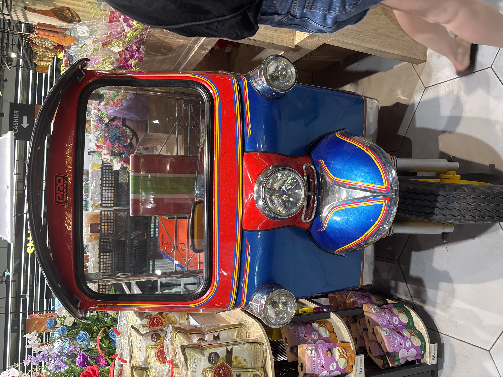 The image shows a tuk-tuk, a three-wheeled vehicle common in Thailand, inside what appears to be a store. The tuk-tuk is decorated in bright colors, primarily red and blue, with yellow accents. It has a small cabin area visible through the front windshield, and the vehicle's front end features several chrome headlights.
The tuk-tuk is surrounded by merchandise, including packaged foods, flowers, and various other items, suggesting it is being used as a display or decorative element within the store.
A sign reading "CASHIER" with an arrow pointing to the left is visible near the top left of the frame.
The environment is a modern store interior with tiled flooring.
In the top portion of the image, a person wearing denim shorts and a dark-colored top is visible. Their legs and feet are also visible, indicating they are standing near the tuk-tuk.
Considering the presence of the tuk-tuk and the Thai language packaging, it is likely the store is located in Thailand. Given the additional textual information about the Thai name "บ้านมะขาม หอบ," which refers to the store, it could be assumed the image was captured in the store "บ้านมะขาม หอบ" in Baan Chaweng, Thailand.