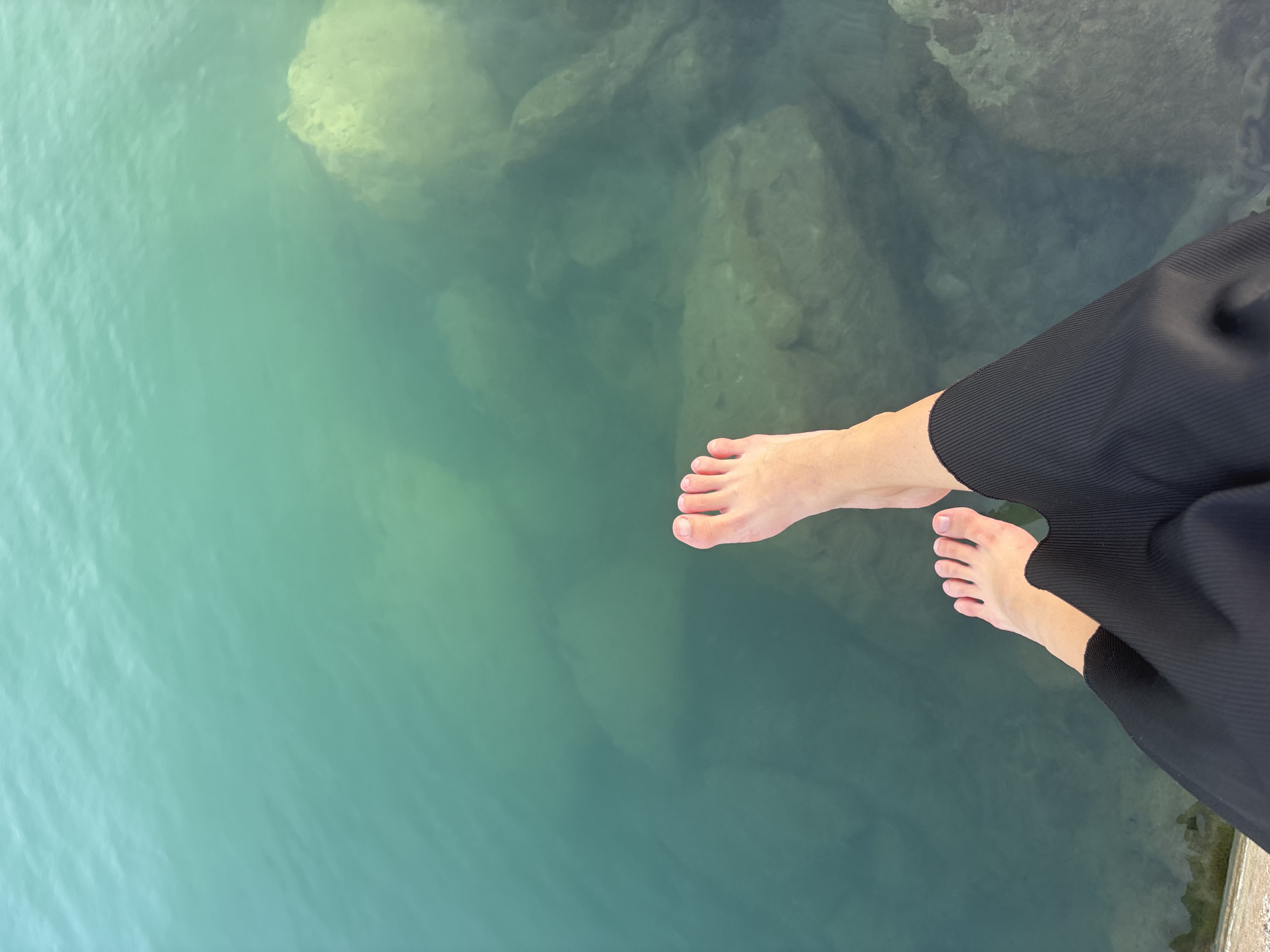 The image shows a person's bare feet and legs, clad in black pants, dangling over the edge of a structure, likely a pier or seawall, in Urbanización Cabo Roig, Spain. The water is a clear, shallow turquoise, revealing submerged rocks and the seabed. The sunlight creates dappled reflections on the water's surface. The perspective is from above, looking down at the feet and into the water.