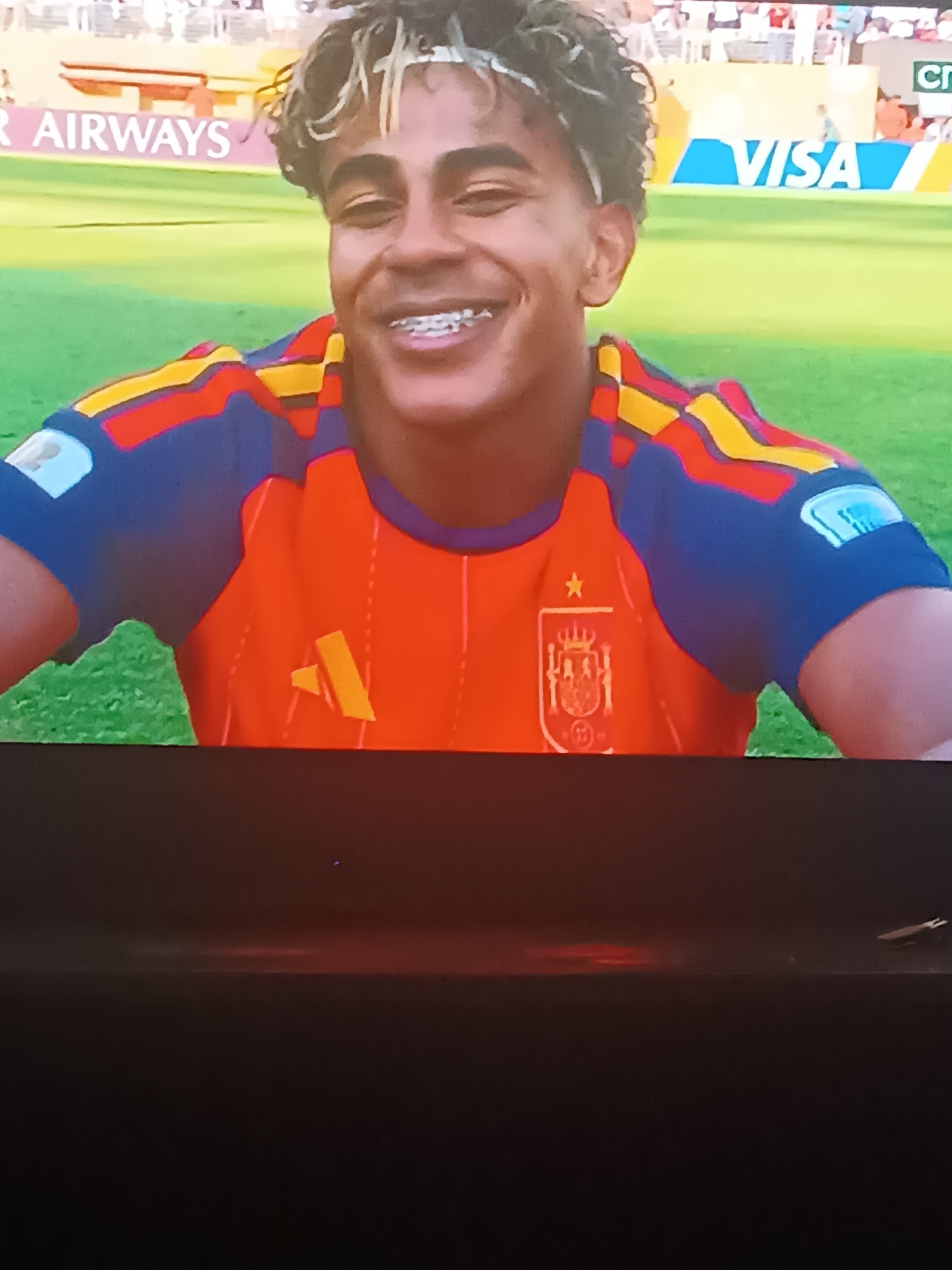 A person with curly, dyed blonde and brown hair is looking at the camera with a wide smile and braces. They are wearing an orange soccer jersey with blue and yellow accents on the shoulders and sleeves. A crest is visible on the left side of the jersey. The background shows a blurred grassy field with advertising banners, including "QATAR AIRWAYS" and "VISA." The image appears to be a screenshot taken from a screen. The location is not discernable from the image.