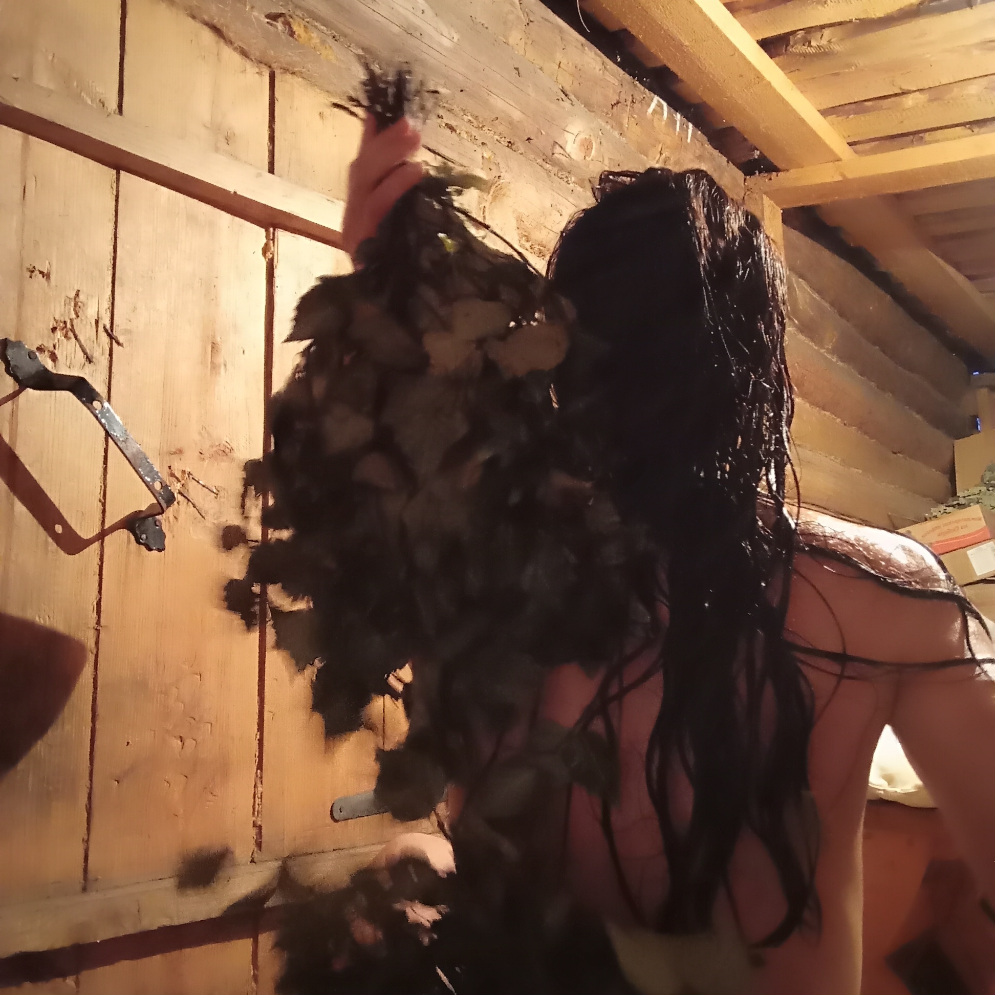 A low-angle shot depicts a person, with long, dark, wet hair, possibly in a sauna, located in the city of Буренка, Russia. The person is holding a bundle of leafy branches. The background consists of wooden paneling on the walls and ceiling. A metal bracket is attached to the left wall. The lighting is dim. Boxes are visible in the background. The person's back and shoulder are partially visible. The person's actions and interactions within the scene are not clearly defined, but it appears they are possibly participating in a sauna ritual.