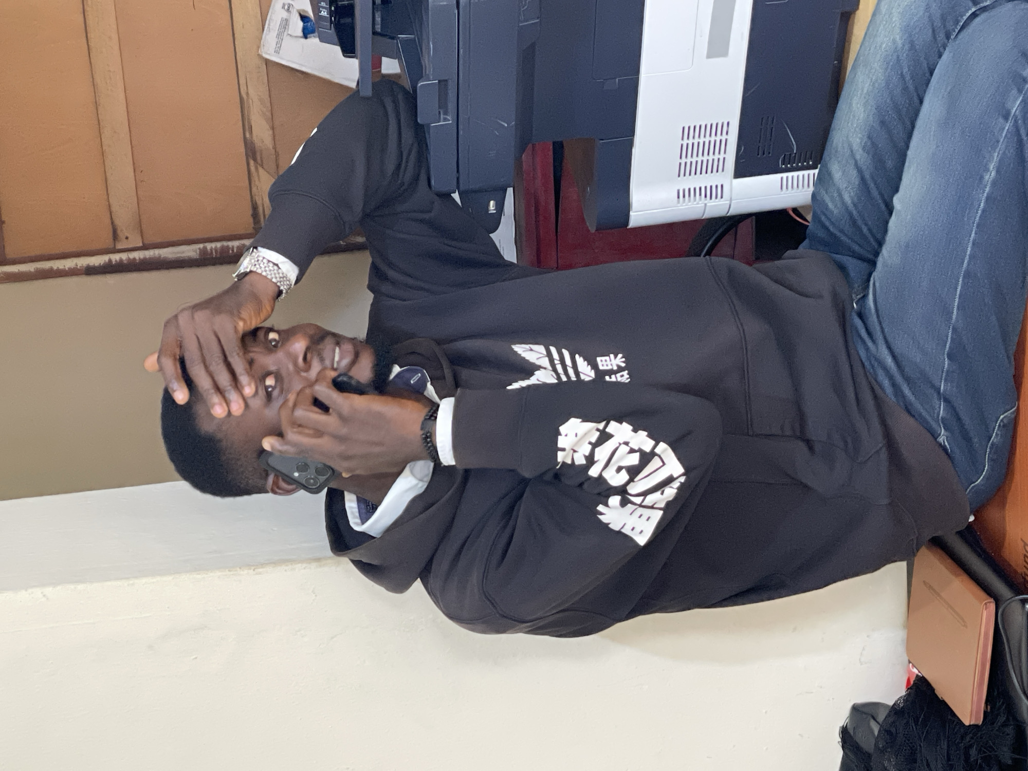 In Durumi, Nigeria, a Black male, appearing to be in his late 20s or early 30s, is seated indoors, looking towards the camera with his right hand touching his forehead. He is wearing a black hooded sweatshirt with white graphic designs on the sleeves and a white collared shirt underneath. He is holding a black smartphone to his left ear with his left hand, engaged in a phone conversation. A silver wristwatch is visible on his right wrist. The background features a wall with a textured finish and wooden planks. To his right, a large black and white printer is present, partially obscuring a dark red filing cabinet. In the foreground, a brown notebook rests on top of a black object. The lighting suggests an indoor environment with overhead light sources.