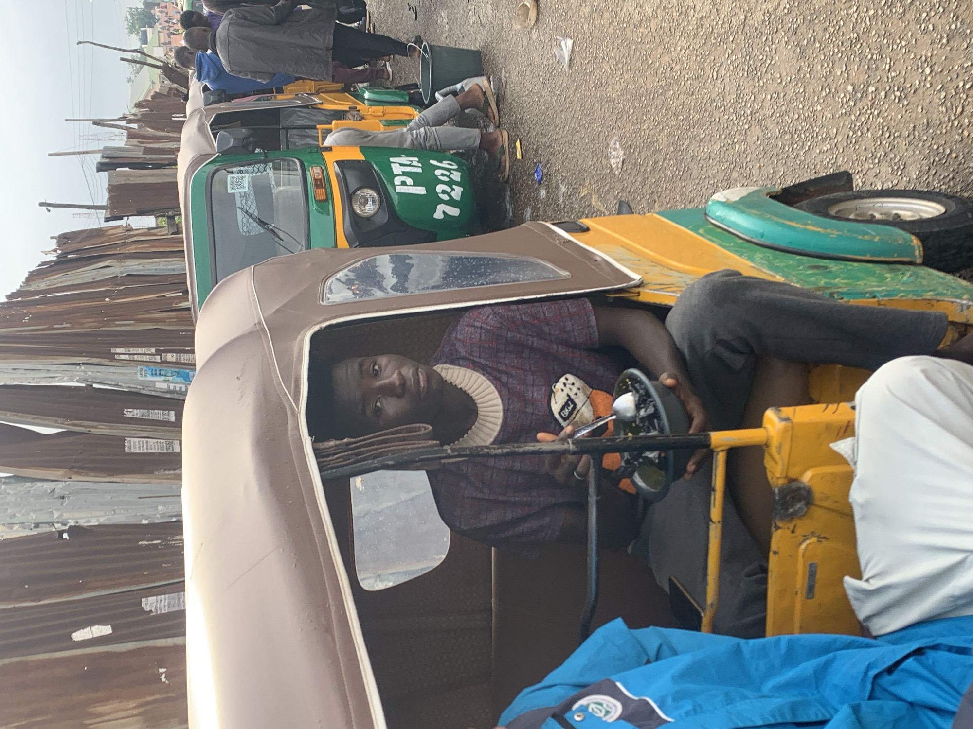 A medium shot captures a young Black male, seated inside the driver's compartment of a yellow and green auto-rickshaw, commonly known as a "Keke Napep," in Jos, Nigeria. He is looking towards the camera with a neutral expression, holding a dark bowl with a spoon, appearing to be eating. The auto-rickshaw's canopy is a muted brown, and the interior shows patterned fabric. A blue garment, possibly a jacket, is draped over the side of the vehicle in the foreground.
Behind the auto-rickshaw, a line of similar vehicles stretches into the distance, suggesting a traffic jam or a queue for passengers. Several other individuals, mostly Black males, are visible standing or sitting around the Keke Napeps. They are dressed in casual clothing, including a blue hoodie and a dark jacket. The background is dominated by a corrugated metal fence, likely a barrier or part of a market stall, with some posters attached. The ground is a rough, asphalt-like surface littered with small pebbles and some scattered debris. The sky is overcast, indicating a cloudy day. The scene appears to be an urban setting in Jos, Nigeria, likely a busy street or transport hub.