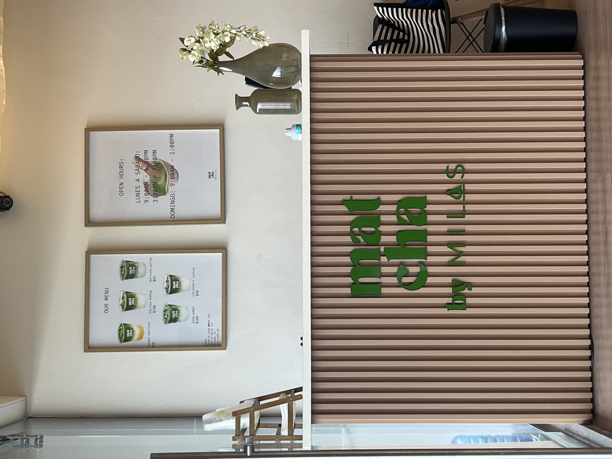 The image shows the interior of "mat cha by MILES" in San Bernardino Tlaxcalancingo, Mexico. On the wall are two framed documents: one is a menu displaying various matcha beverages and their prices (Mango Matcha - $90, Salted Maple - $100, Matcha Latte - $85, Coconut Cloud - $90, and Earl Grey - $100), the other is a list of opening hours (Monday to Saturday 9:00 AM - 1:00 PM and 3:00 PM - 7:00 PM, Sunday 9:00 AM - 1:00 PM). To the right of the framed documents is a light-wood panel with the words "mat cha by MILES" in large green letters. On top of the wood panel are various decorative items: a vase containing white flowers, a translucent green container, and a small bottle of what appears to be cleaning solution. There is also a black-and-white striped tote bag and black cylindrical container. In the lower portion of the frame, there is a container of disposable cups.