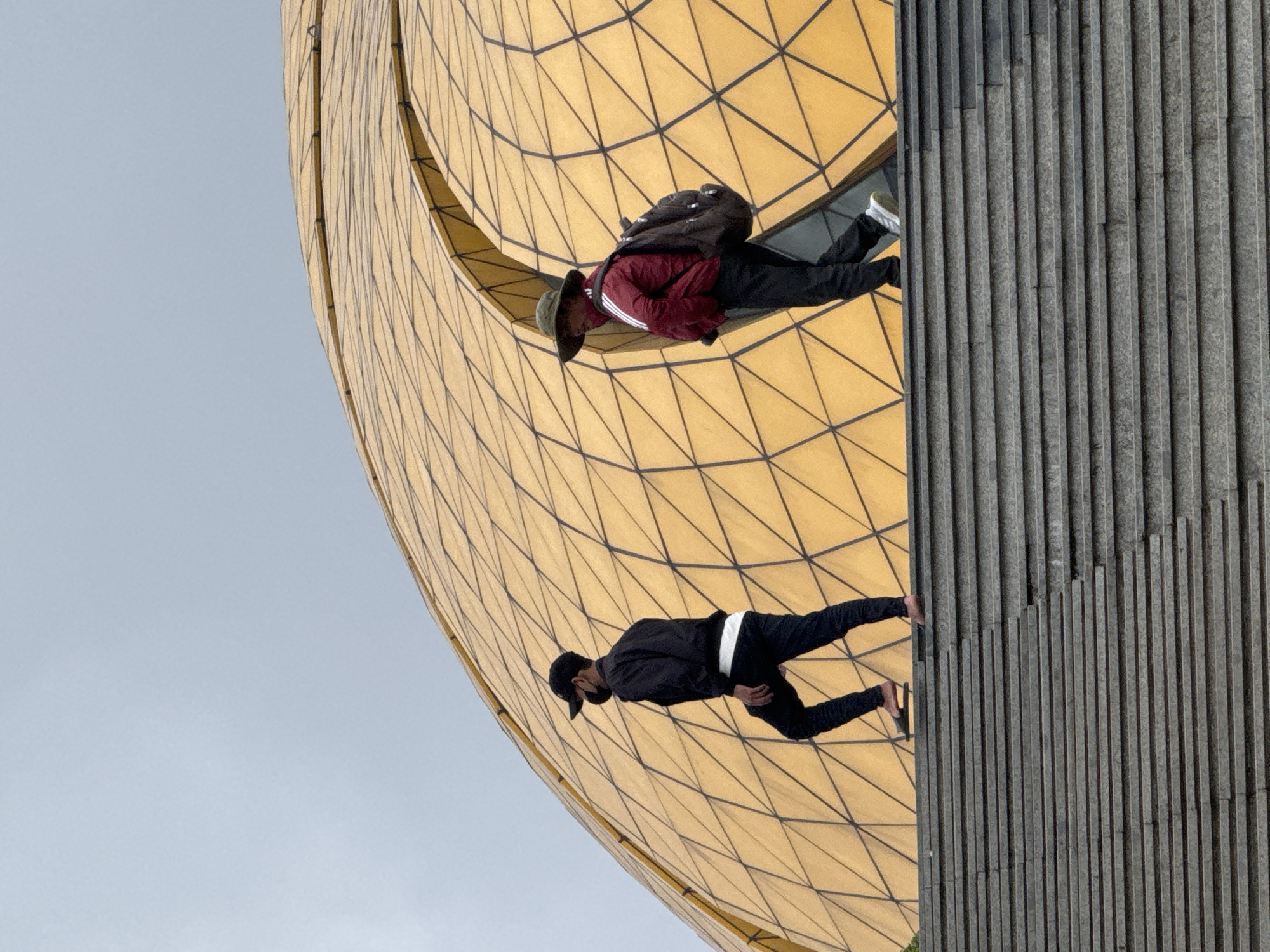 This media file presents a photograph intentionally rotated ninety degrees counter-clockwise, creating an optical illusion of people walking on a curved wall. The prominent feature is a large, golden-toned architectural structure with a tessellated, geometric grid pattern, forming a vast, concave surface that dominates the left and central parts of the image. To the right, a vertical section of a grey, ribbed wall or paneling is visible. A pale, light blue-grey sky occupies the background, visible on the left side of the image.
Two individuals are depicted on the golden structure, appearing to defy gravity. The person positioned more centrally wears a dark baseball cap, a black jacket, black pants with a white stripe at the waist, and dark footwear. They are shown mid-stride, facing right. Further to the right, a second person wears a dark, wide-brimmed hat, a maroon jacket with white stripes on the sleeves, black pants, and white sneakers, carrying a dark backpack. This individual is also in a walking or posing stance, appearing to step onto the grey wall from the golden one.
The scene is illuminated by natural daylight, indicated by the soft, even light and the light sky, suggesting it is either an overcast day or the sun is not directly overhead. The unique, modern architecture, particularly the golden geodesic-like structure, serves as a distinctive location cue within Da Lat, Vietnam, where the photo was taken. The subjects are likely posing for a creative photograph, leveraging the architectural design and the rotated perspective. No visible text is present in the image.