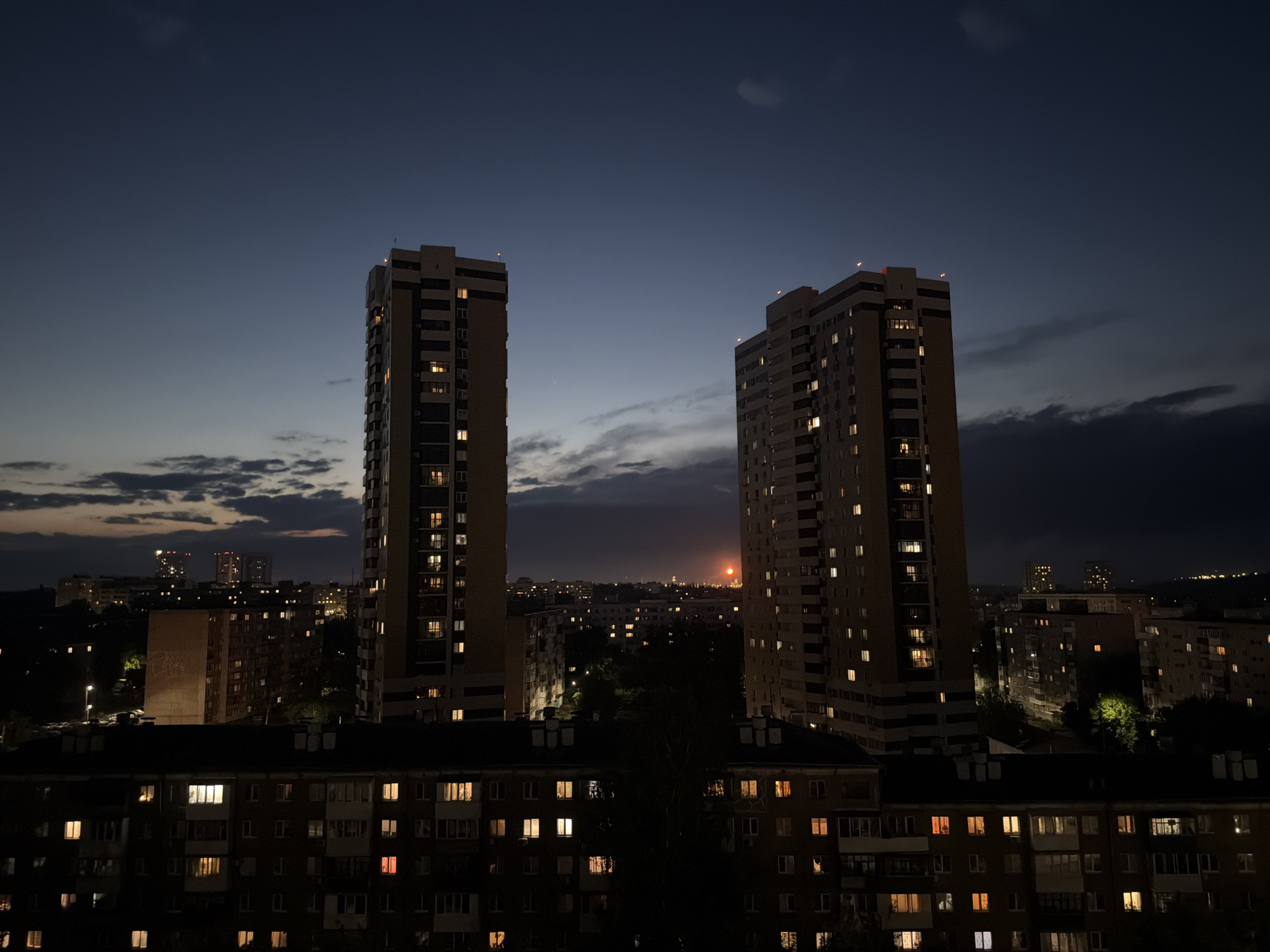 The image shows a nighttime cityscape in an unknown city. Two tall, beige-colored apartment buildings dominate the foreground, with lights visible in many of their windows. Below these buildings, a shorter, darker apartment building is seen with numerous illuminated windows. In the background, other buildings and a city skyline are faintly visible under a dark blue and cloudy sky, with a small orange light in the distance. There are no people visible in the image. The environment is urban and residential.