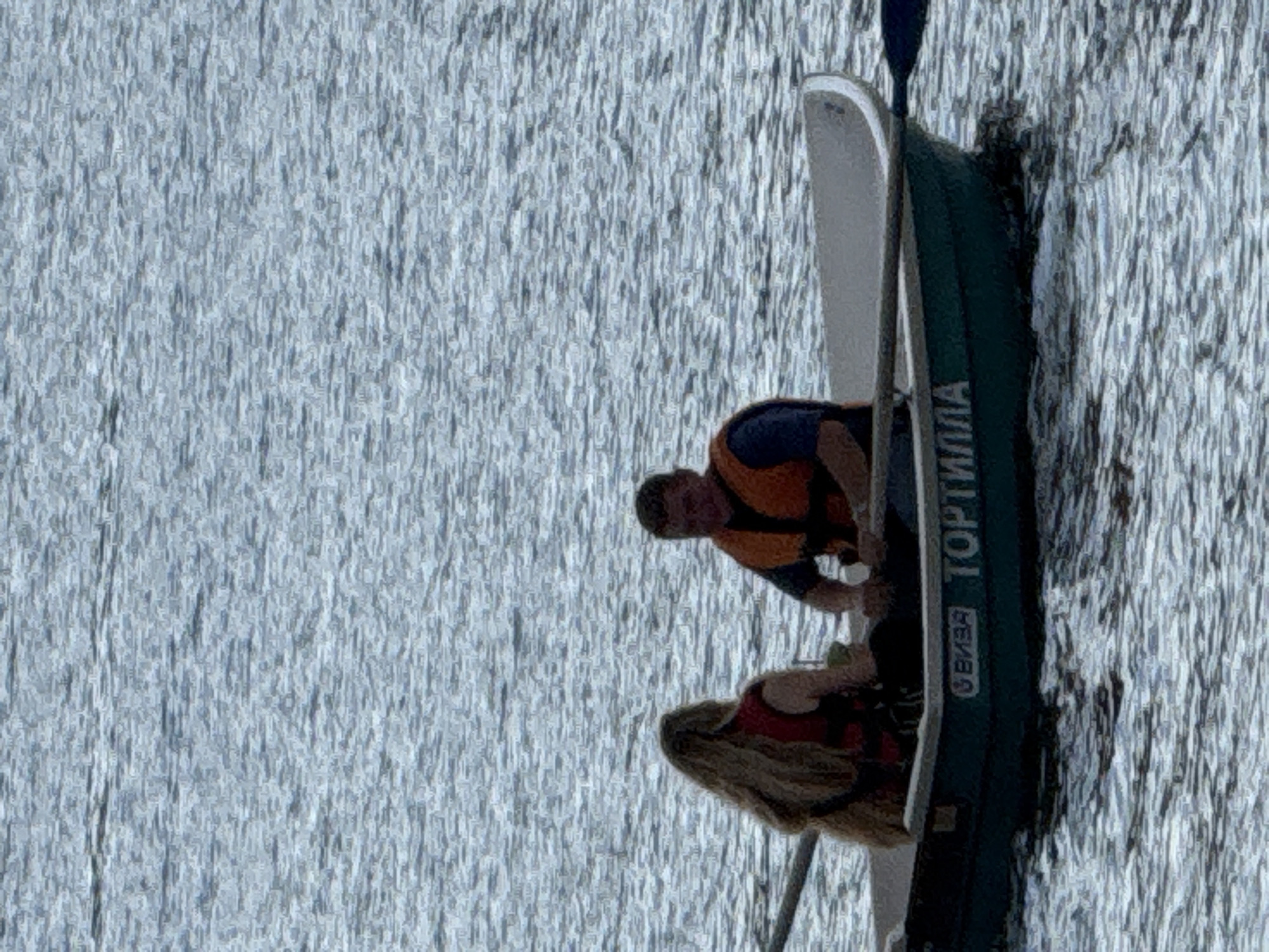 Two individuals, a man and a woman, are seated in a small green boat on a body of water. The woman, facing away from the camera, has long blonde hair and is wearing a red life vest. The man, facing the camera, is wearing an orange life vest and appears to be holding something. The boat has text on its side that reads "ТОРТИЛЛА". This scene is on the Iset River in Yekaterinburg, Russia, near the dam of the Yekaterinburg Ice Palace. The water is choppy with visible ripples.