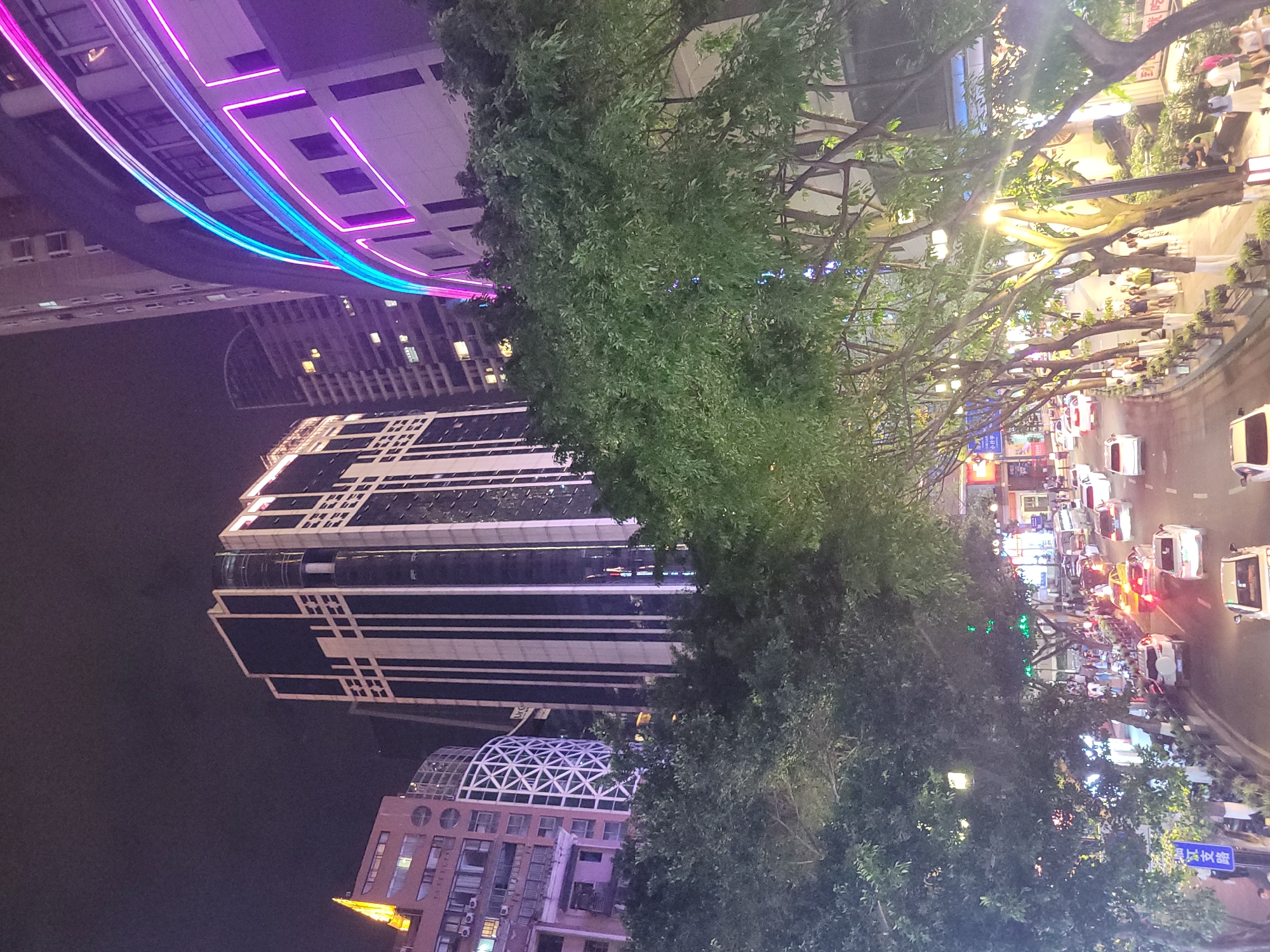 This image captures a vibrant night scene in Yuzhong District, China, showcasing a bustling urban environment. The perspective is from an elevated position, looking down upon a street flanked by buildings and trees.
On the left side of the frame, several modern architectural structures dominate the skyline. A tall building features distinctive dark and light geometric patterns, while a lower building has a unique geodesic dome-like roof. In the upper left, a large building is spectacularly lit with prominent pink and blue neon strip lights, creating a dynamic, arcing design that reflects brightly.
A dense line of leafy green trees runs through the middle ground, effectively separating the towering buildings from the active street life. These trees are partially illuminated by the myriad of artificial lights from the surroundings.
The right side of the image reveals a busy city street and sidewalk. Numerous people are visible, walking, congregating, and possibly dining at outdoor establishments. The street is active with various vehicles, including cars and motorbikes, their headlights and taillights creating streaks of light in the darkness. The storefronts lining the street glow with a multitude of bright commercial signs and streetlights, contributing to the energetic atmosphere.
The time of day is clearly night, as indicated by the dark sky and the pervasive use of artificial lighting. The weather appears clear. A notable detail is a blue street sign on the right side, which clearly displays Chinese characters, reading "望龙门" (Wanglongmen), providing a specific location cue within Yuzhong District. The overall impression is one of a lively and active urban center after dark.