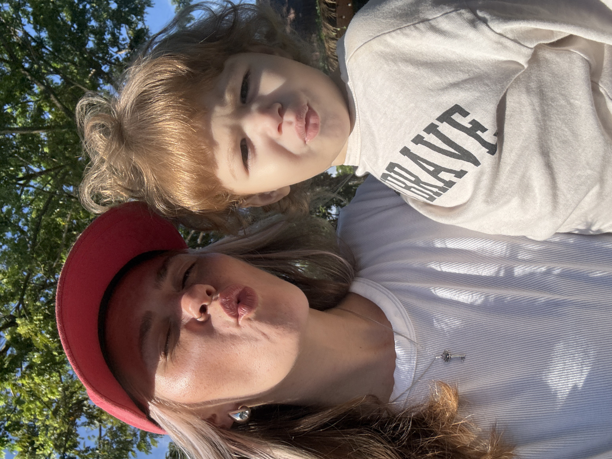 A close-up, front-facing selfie captures a woman and a young child outdoors. The woman, wearing a red baseball cap and a white, ribbed t-shirt with a silver key pendant, puckers her lips as if for a kiss. She has blonde hair and is wearing a silver hoop earring. Beside her, the child, with light brown curly hair, also puckers their lips. The child is wearing a light-colored long-sleeved shirt with the word "BRAVE" printed in dark, block letters across the chest. Sunlight casts shadows on their faces and clothing. Lush green trees and a bright blue sky form the background. The setting appears to be outdoors in a warm climate, potentially a park or garden, within the locale of Choeng Thale, Thailand.