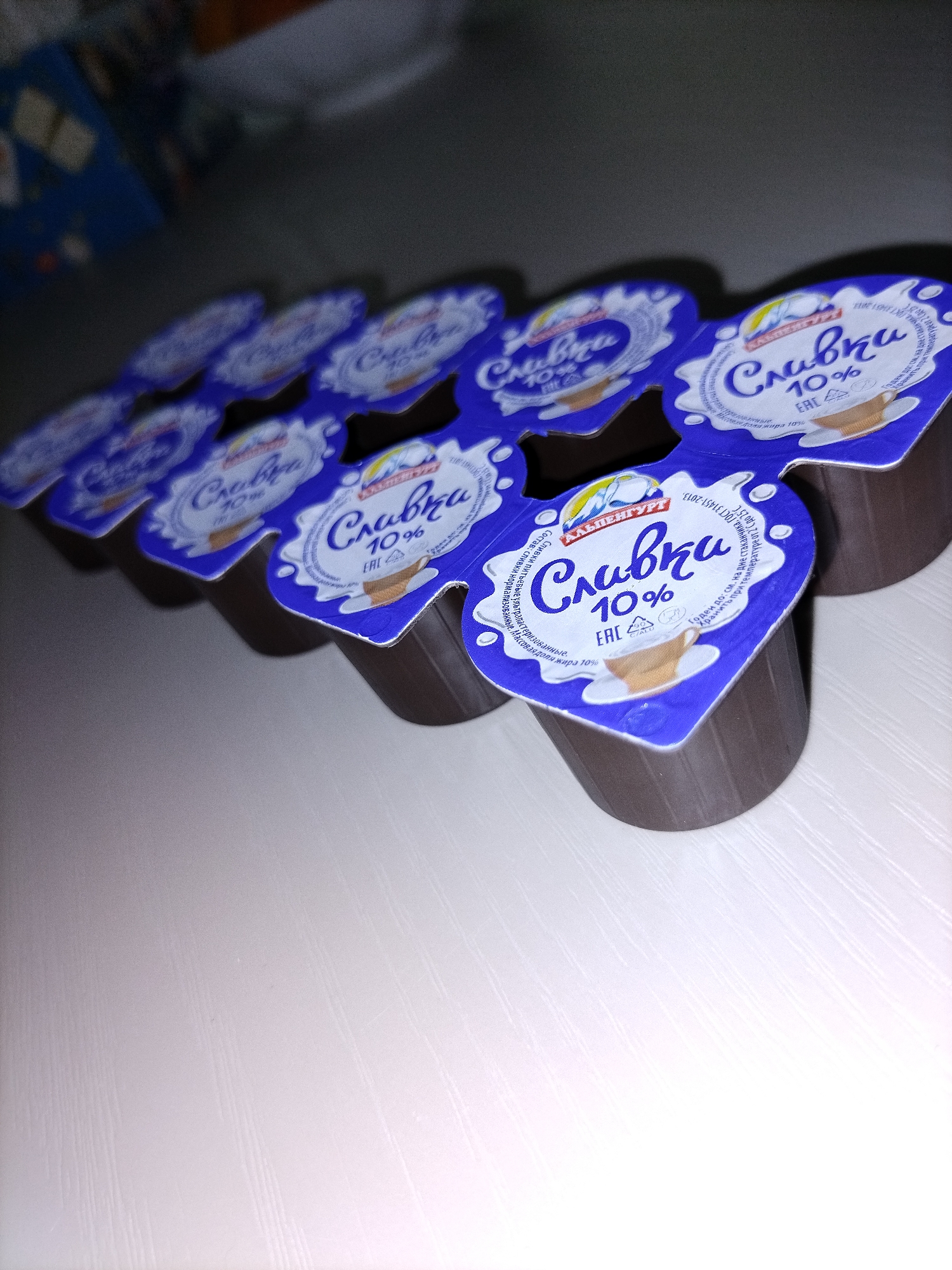 The image shows a close-up, angled shot of multiple small, individually packaged servings of dairy cream, likely creamers for coffee. These are arranged in a staggered, fan-like pattern on a light-colored, textured surface, possibly a table. Each creamer has a blue lid with white lettering and graphics. The dominant text on the lids reads "Сливки 10%" (Slivki 10%), indicating the product is 10% fat cream. Some lids also feature images of a teacup with cream and a sun-like emblem. The packaging appears to be made of plastic, with ribbed sides visible on the creamer cups. The background is blurred and dark, with hints of what might be packaging or other items. The lighting suggests an indoor setting. The scene contains no people, and the objects are static. There is no identifiable indication of a specific location within the city of Смалявічы, Belarus, as the image focuses solely on the product packaging.