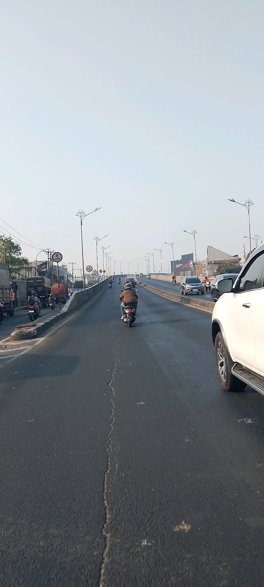The image captures a view from a moving vehicle on an elevated road in Bekasi, Indonesia. The road is filled with traffic, predominantly motorcycles, with a few cars and trucks visible. A lone motorcyclist in a brown jacket is in the foreground, heading away from the camera. On the left side of the road, there are several trucks and other vehicles. Streetlights line the elevated road, and the sky is clear and bright, suggesting it is daytime. The infrastructure includes concrete barriers and signage, including a speed limit sign of 40. The road surface shows signs of wear and tear with visible cracks. The overall scene depicts a typical urban traffic environment in Bekasi.