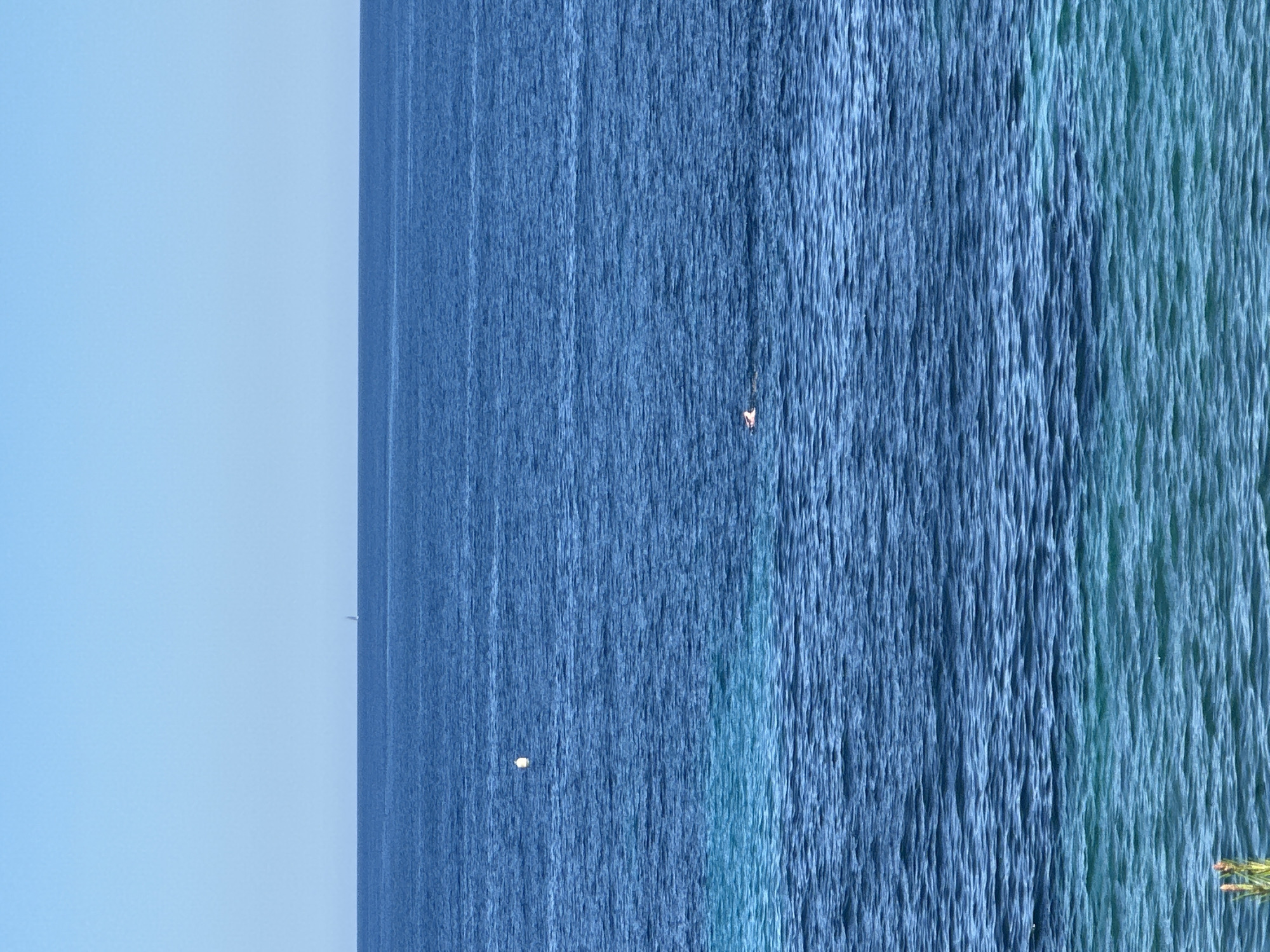 The image depicts a vertical shot of the sea along the coast of Teulada, Spain. The sea transitions from dark blue to turquoise and the sky is visible on the left side of the frame, appearing a clear, light blue. Small objects, possibly boats or birds, are visible in the distance on the sea.