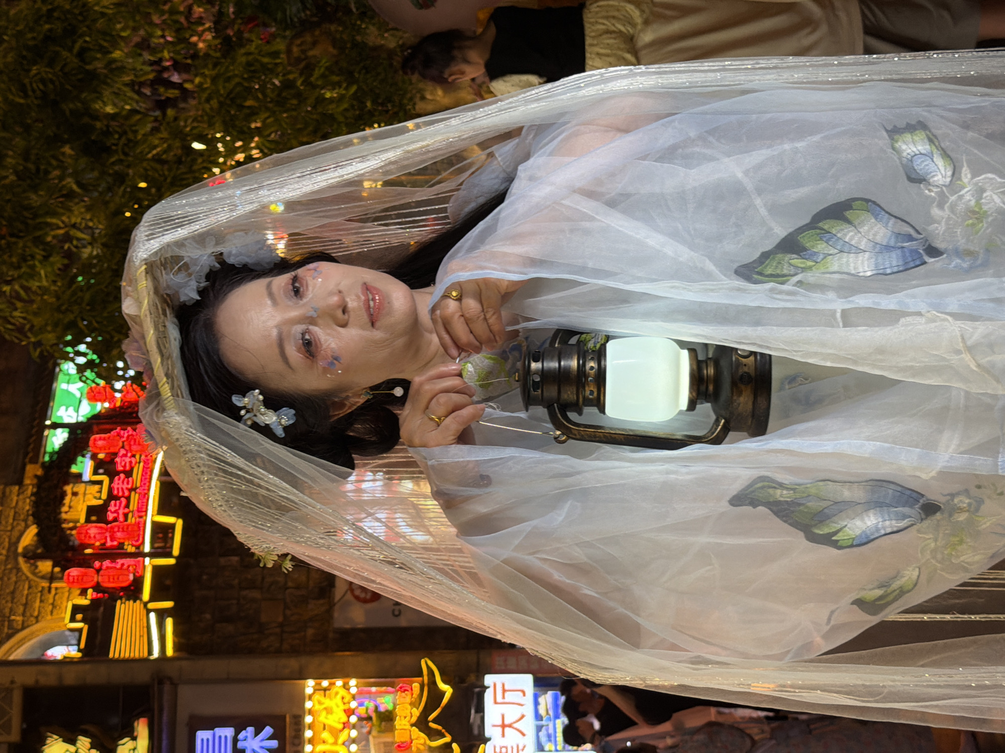In Yuzhong District, China, a woman with a veiled headdress and traditional attire stands holding a glowing lantern. Her makeup includes glittery accents on her face and eyes. The background is a street scene at night, illuminated by bright neon signs and decorative lights. The woman's white, sheer outer garment features embroidered butterflies and floral patterns. In the distance, other people and a child are visible, suggesting a public gathering or event.