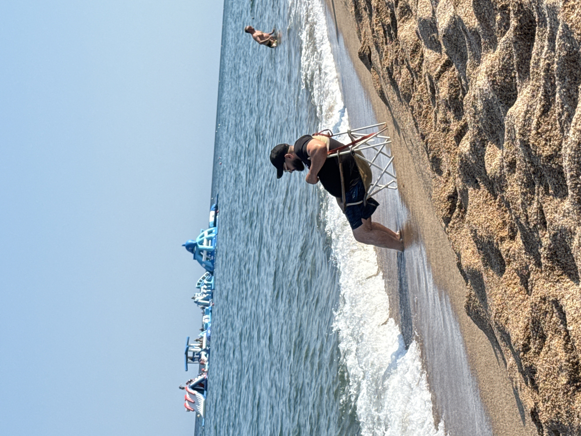 The image captures a sunny day at a beach in Blanes, Spain. The scene is dominated by a sandy shore with textured dunes on the right and the shimmering blue ocean on the left. A man wearing a black t-shirt, black shorts, a black baseball cap, and sunglasses is the most prominent subject. He is leaning forward, seemingly holding onto a light-colored, open-frame chair. His pose suggests he might be looking at something in the water or preparing to sit.
Further out in the ocean, another person, a man, is visible lying in the water. In the background, along the waterline, there are several inflatable structures, likely part of a water park, characterized by their bright blue and white colors. The ocean appears calm with gentle waves lapping the shore. The sky is clear and a bright blue, indicating fair weather and likely midday. The overall atmosphere is one of leisure and recreation. The sand is coarse and appears dry in some areas, with wet patches closer to the water. The lighting suggests it is daytime, with shadows cast by the dunes.
