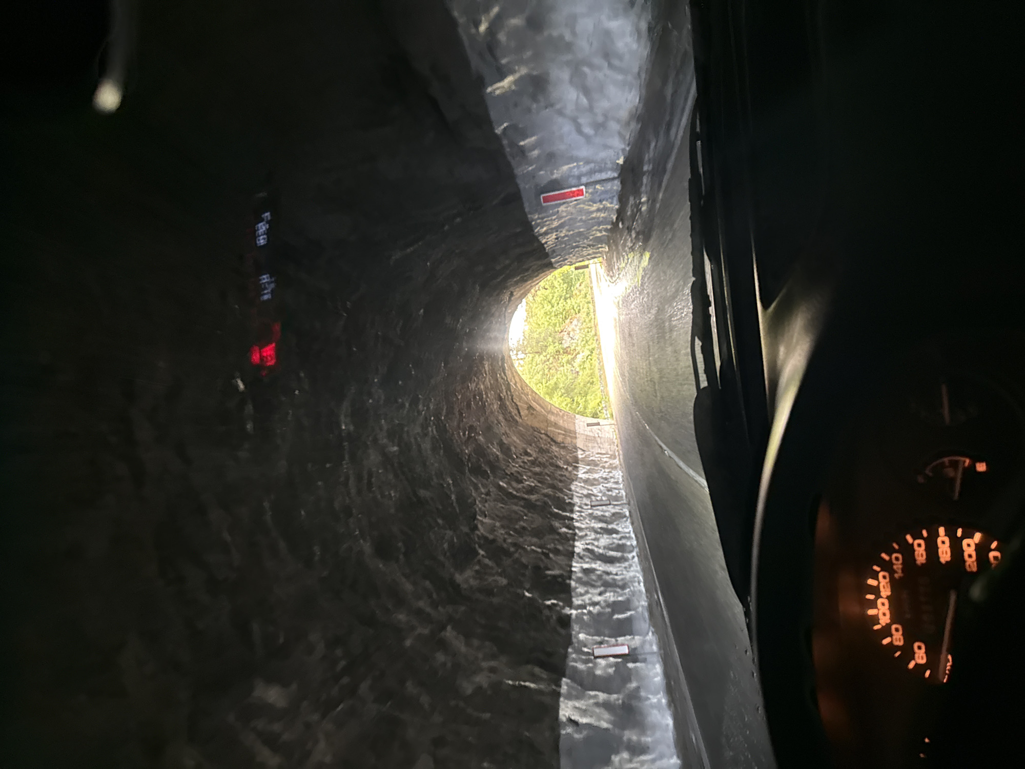 The image depicts the view from inside a vehicle traveling through a long, concrete tunnel, possibly a drainage tunnel or culvert. The tunnel walls are uneven and textured, with small, rectangular markers placed along the sides. The end of the tunnel shows an opening to the outside, revealing green foliage and daylight. Inside the vehicle, part of the dashboard and speedometer are visible, illuminated in orange light. The location is likely Колишовци, Bulgaria, implied from the prompt. There is no apparent human activity in the image.