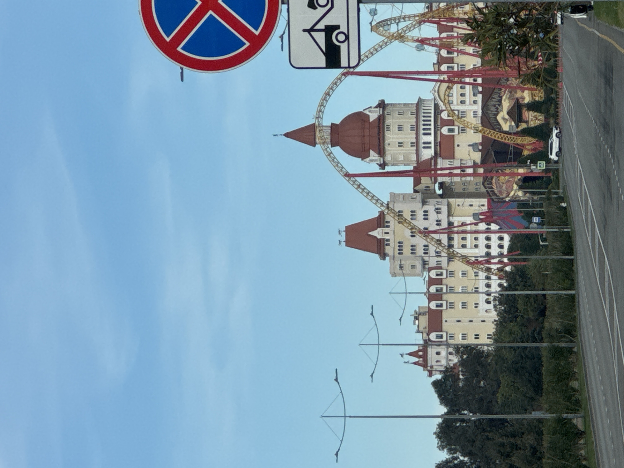 The image shows a large building with reddish-brown roofs and white walls, featuring multiple towers and windows. A roller coaster is visible behind the building with red supports and golden tracks. The building is likely in Sochi, Russia, given its architectural style. A road runs along the right side of the image with parked cars. Streetlights line the road between the building and the road. In the upper left are road signs: a parking prohibition sign and a parking sign for disabled vehicles. The sky is a light blue. The image is oriented sideways.