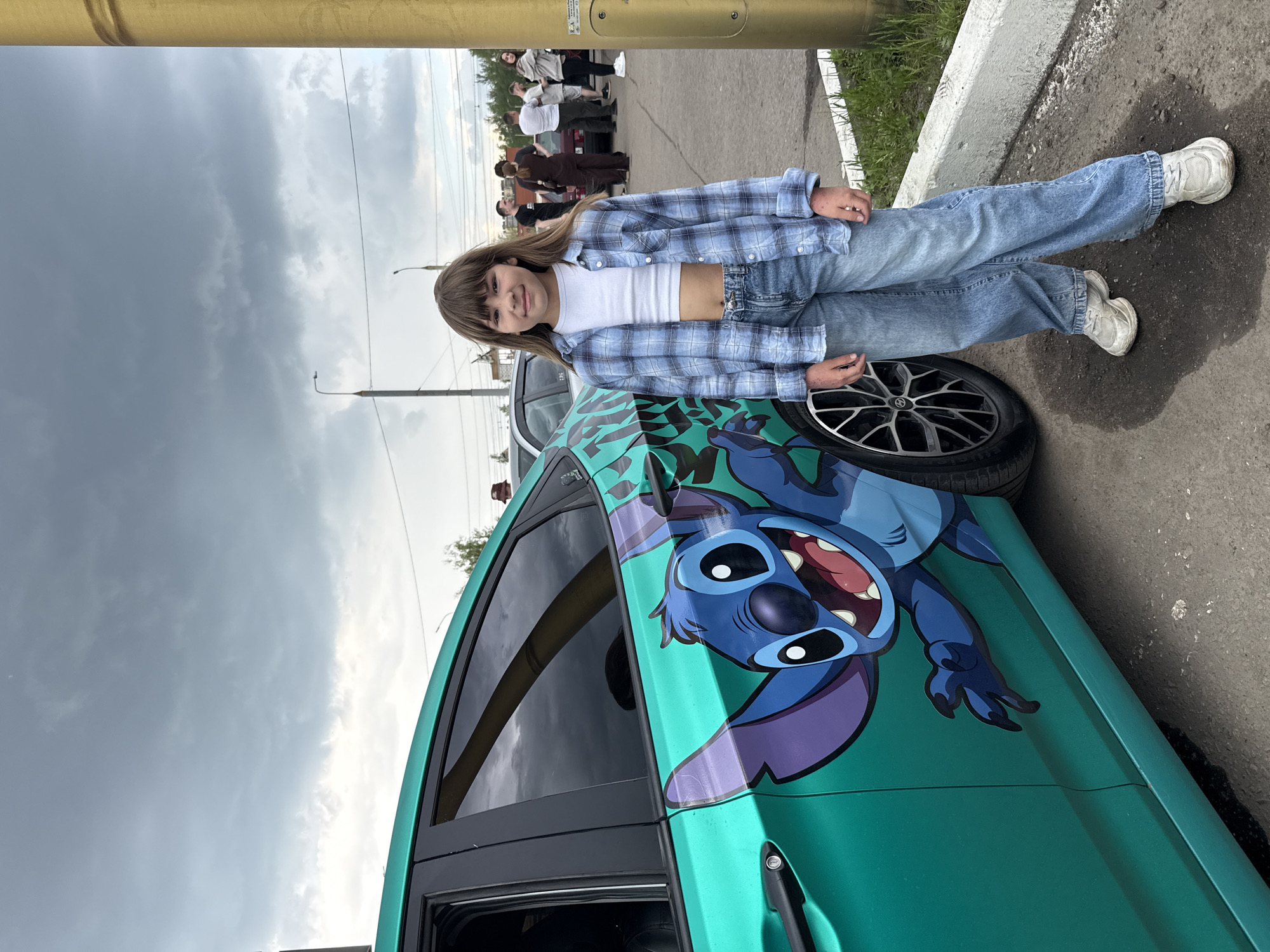 A young girl leans on a car with a large, colorful cartoon character (Stitch from "Lilo & Stitch") painted on its side. She wears a white crop top, blue plaid shirt, and jeans, with white sneakers. The car has a spare tire next to it. In the background are other people and a partly cloudy sky. The location is in the city of Rusanovo, Russia.