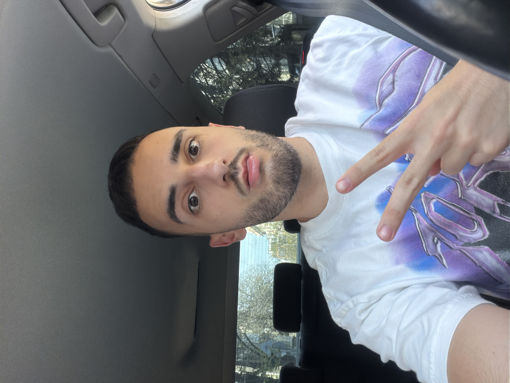A young man is inside a car, making a peace sign with his right hand. He has short, dark hair, dark eyes, and a beard. He is wearing a white t-shirt with a purple and gray design. The interior of the car is gray. The background shows trees and buildings outside the car windows. It is difficult to ascertain if this image was taken in the city of Plovdiv, Bulgaria, because there are no specific landmark buildings, street signs, or other indicators in the photo.