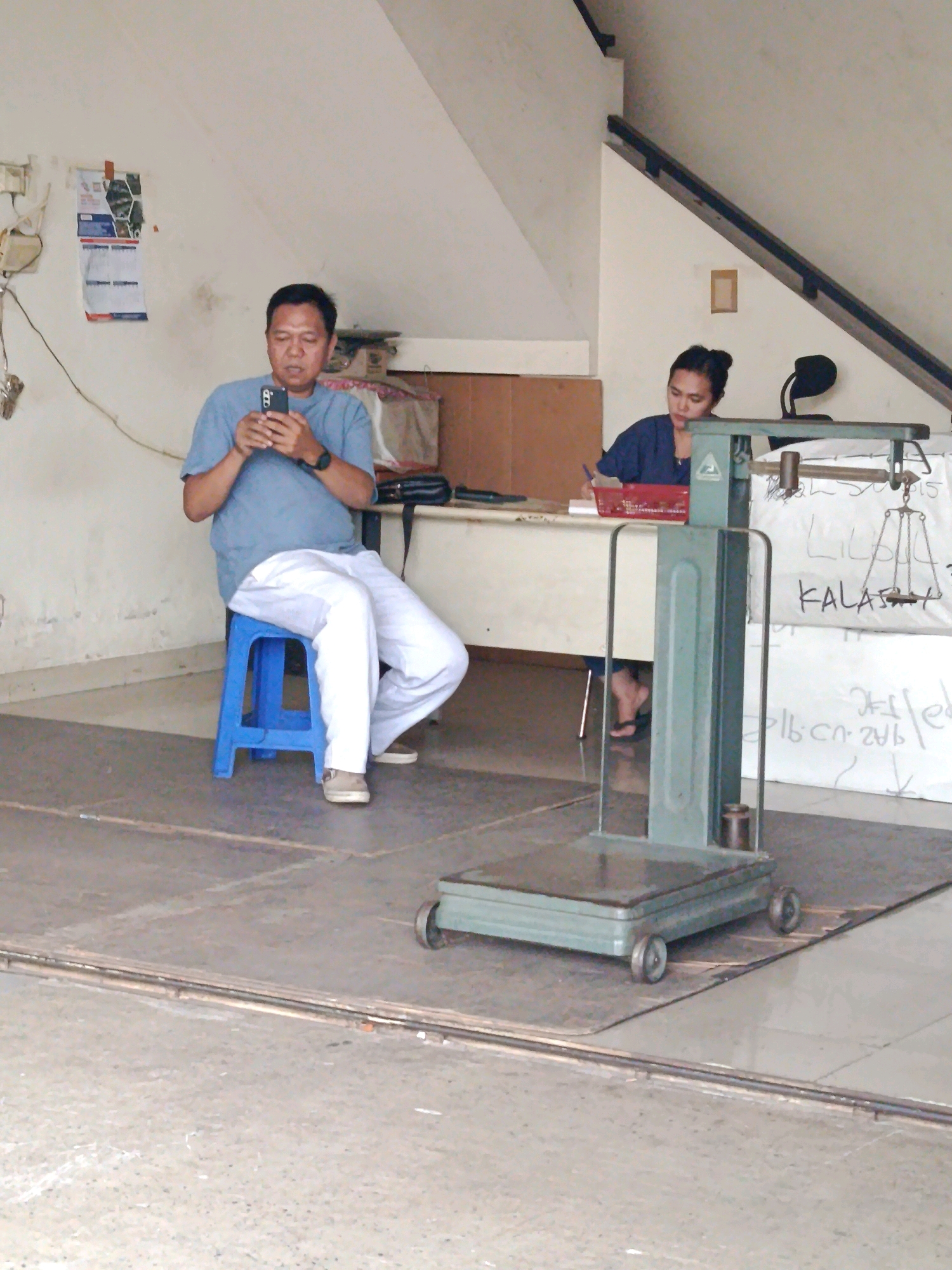 An overhead, slightly angled medium shot captures a scene within a dimly lit, interior space that appears to be a shop or a warehouse in Unknown, Indonesia. The floor is a tiled surface. In the foreground, a green industrial scale with wheels is positioned on the floor.
To the left, a man wearing a light blue t-shirt and white pants sits on a bright blue stool. He is looking down at a black smartphone in his hands. To his right, a woman in a dark blue top is seated behind a desk or counter, looking down and writing on a piece of paper with a pen. A red basket is on the desk in front of her. Behind her, a scale is visible, and a white sheet with handwritten text is pinned to the wall. A black lamp is also visible on a shelf behind the woman. The background wall is a light color, and a staircase with a dark railing is visible on the upper right. There are also some boxes and hanging items in the background. The overall lighting is subdued.