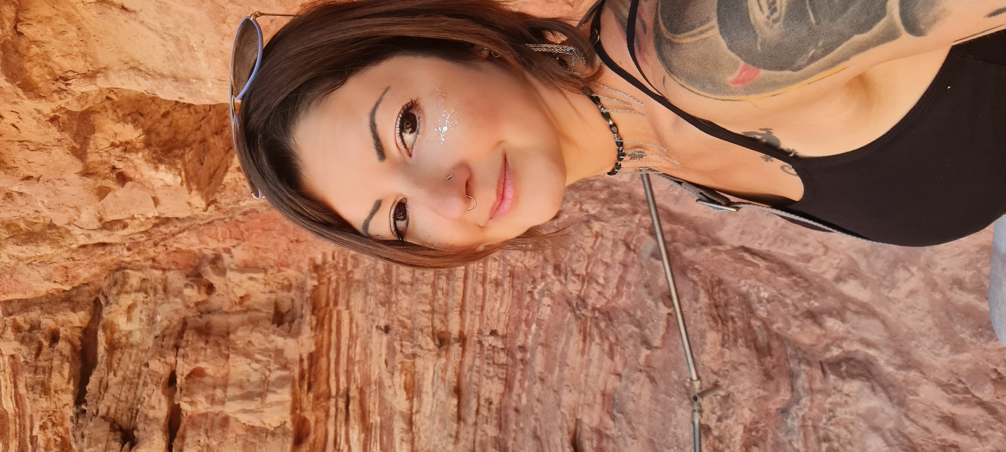 A woman with shoulder-length brown hair stands in front of a geological formation of orange and brown rock. She wears a black tank top and has multiple tattoos visible on her chest and shoulder. Sunglasses are propped on her head. She has glitter makeup around her eyes and a nose piercing. She wears a beaded necklace with metal chains hanging from it. The background is composed of the striated rock formation. The location is likely in the Hevel Eilot Regional Council, Israel, due to the rock formation resembling those in the region.