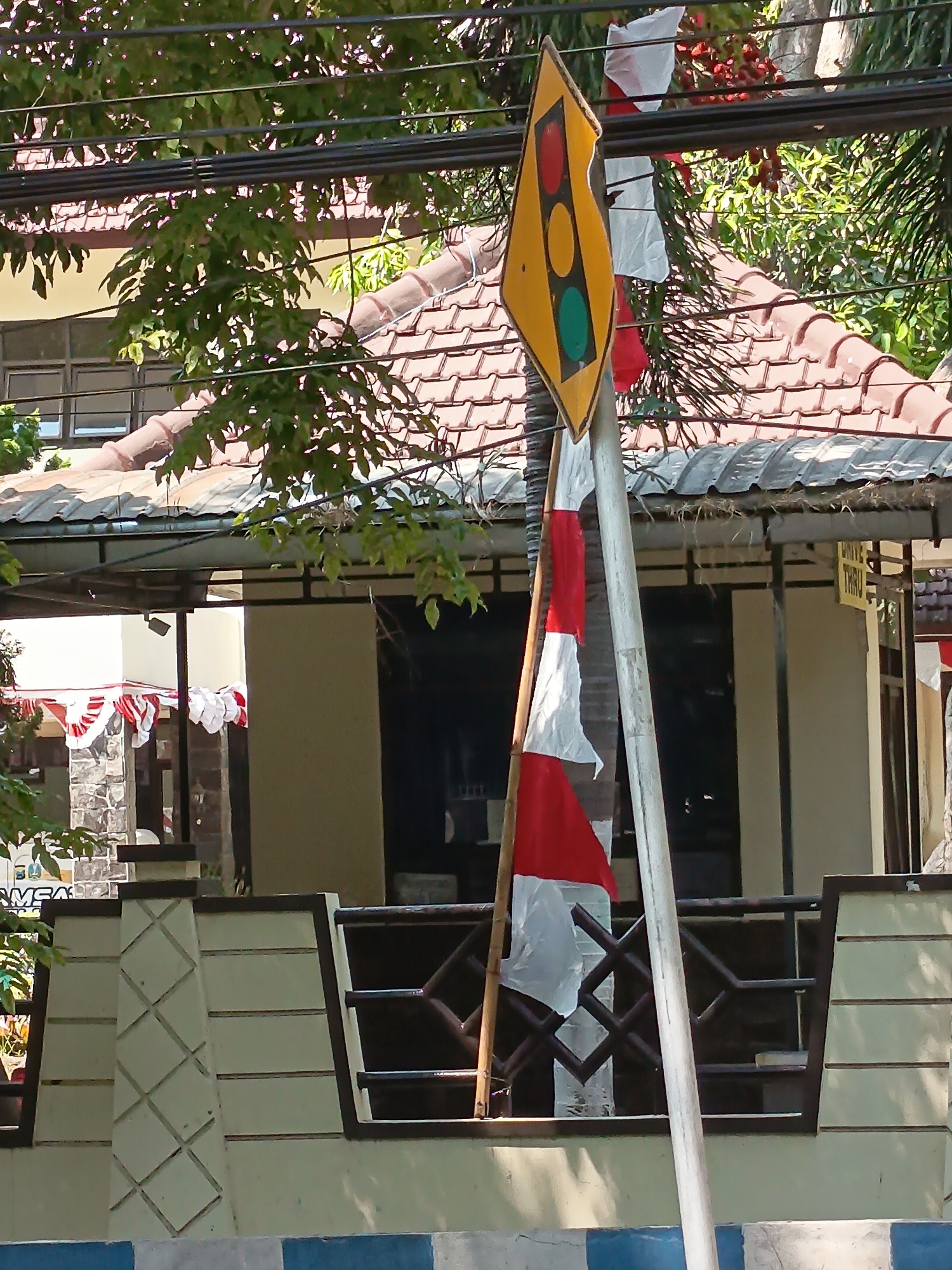 The image captures a street scene in Kandangjati Kulon, Indonesia, on a sunny day. Dominating the foreground is a traffic sign shaped like a diamond, colored yellow with a depiction of traffic lights showing red, yellow, and green. The sign is attached to a grey metal pole, which is partially obscured by a bamboo pole supporting an Indonesian flag. The flag, with its red and white horizontal stripes, is draped around the bamboo pole. Behind the flag and sign, a building with a terracotta tiled roof is visible. A fence with a patterned railing separates the foreground from the building. Trees with green foliage are interspersed throughout the scene, casting shadows on the ground. Overhead, power lines run across the frame. The environment appears to be a typical urban or suburban setting with a residential or commercial building.