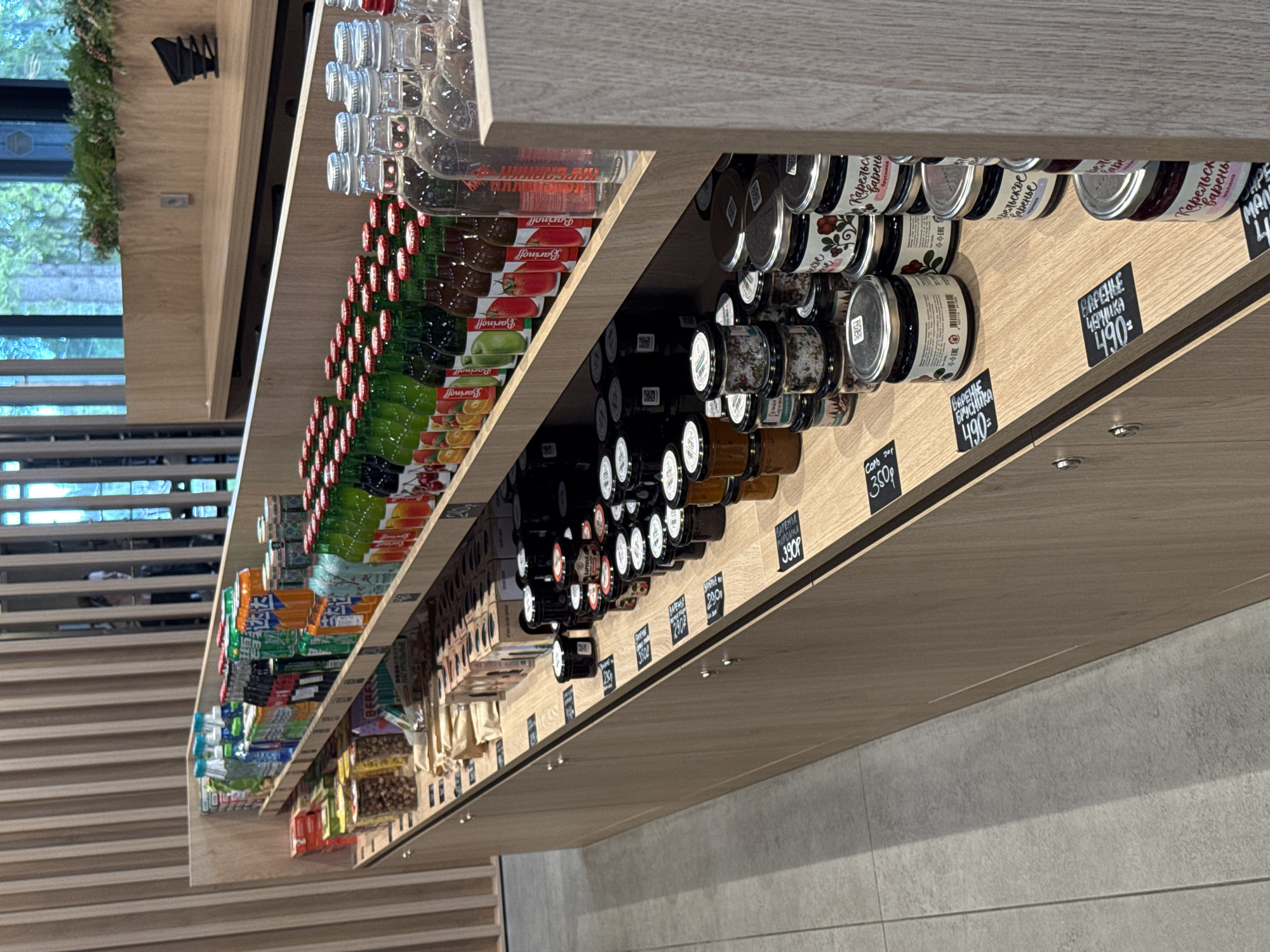 The image shows a retail display shelf stocked with various food and beverage items. The shelf is made of light-colored wood with vertical wooden slats on the back wall. On the top shelf, there are rows of bottled drinks including soda and juice, packaged snacks, and what appear to be jars of preserves or sauces. The lower shelves are predominantly filled with glass jars of preserves, many with labels indicating fruit flavors like cherry, raspberry, and cranberry. Some jars have chalk-like price tags in front of them. The overall environment suggests a small grocery store or a specialized food shop. The lighting is bright, indicating an indoor setting.