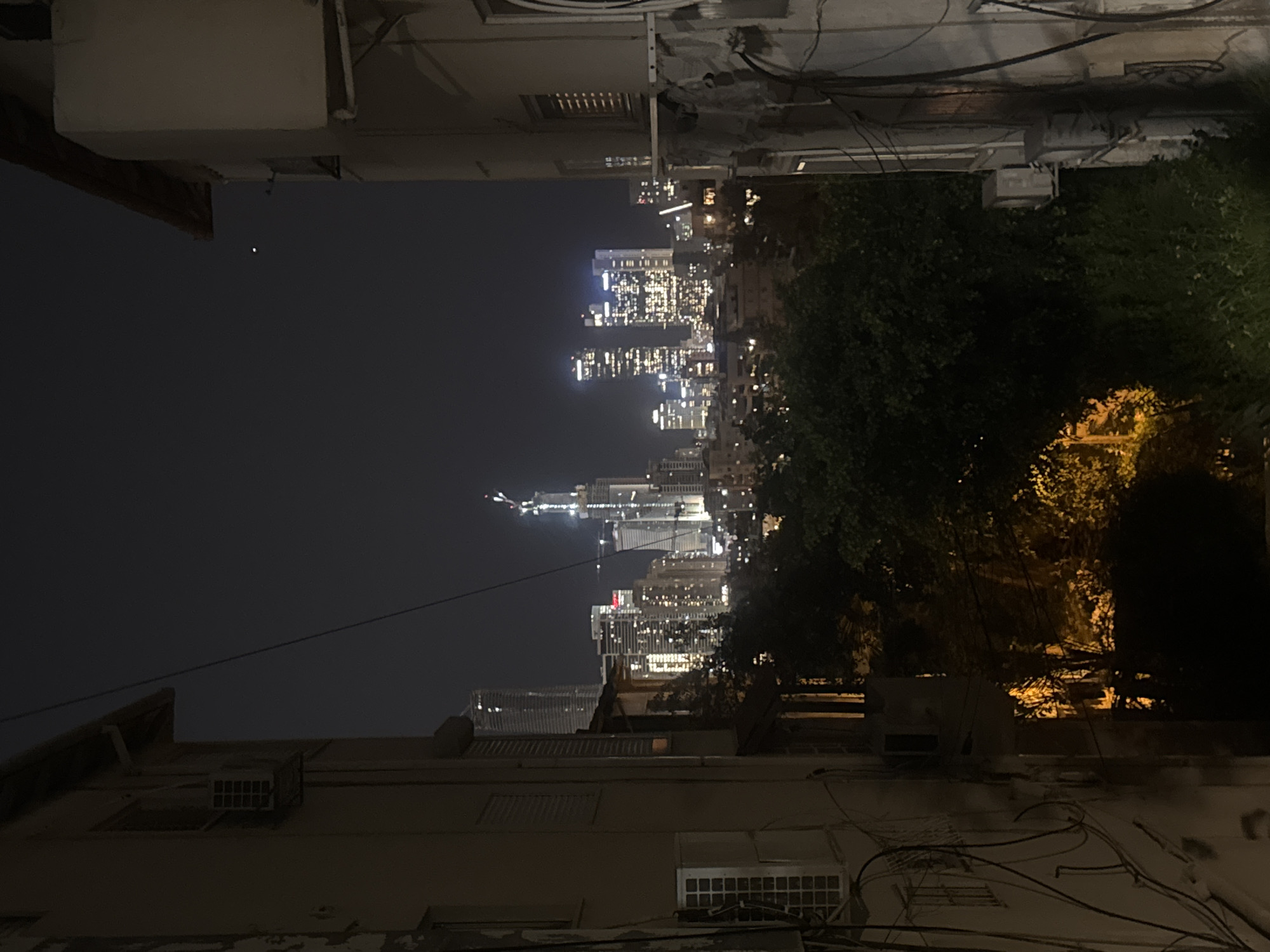 A nighttime view from Givatayim, Israel, looking towards the city center. Illuminated skyscrapers rise in the distance, interspersed with dark patches possibly indicating lower buildings or residential areas. Dark foliage and buildings are visible in the foreground. Overhead wires stretch across the scene. An unknown point light is visible in the sky.