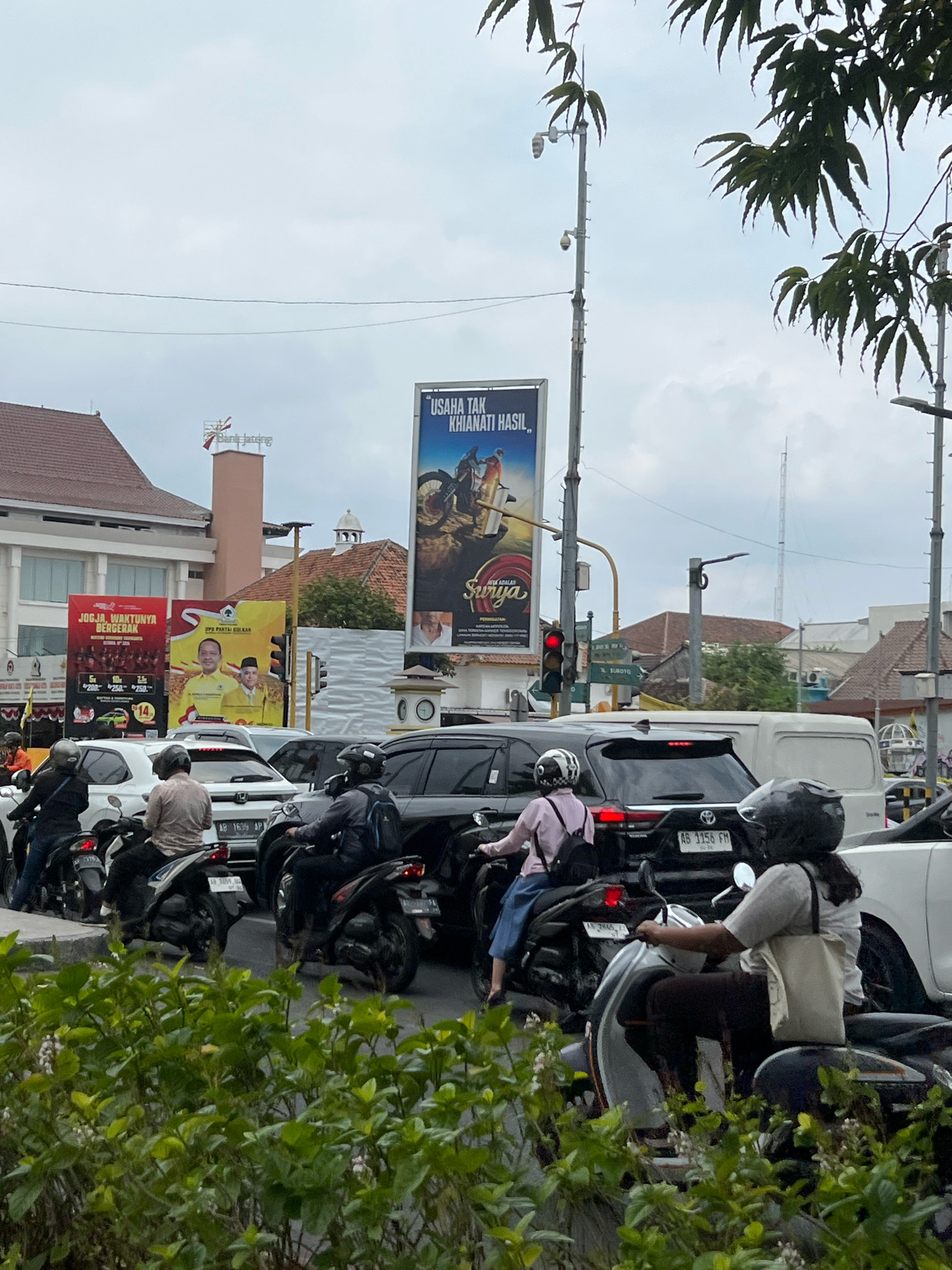The image depicts a street scene in Yogyakarta, Indonesia, characterized by heavy traffic congestion. Several motorcycles, cars, and a van are visible on the road, suggesting a busy urban environment. The foreground is dominated by lush green foliage, partially obscuring the view of the road and vehicles.
In the background, various buildings and advertisements contribute to the city's landscape. A prominent billboard features an advertisement with text that appears to be in Indonesian, alongside images of people and vehicles. Another billboard displays political imagery, with portraits of men and text related to "DPR PARTAI GOLKAR." The presence of traffic lights indicates an intersection or busy junction. The sky is overcast, with clouds creating a diffused light. The overall impression is one of a typical day in a bustling Indonesian city, with the natural element of greenery providing a contrast to the man-made structures and urban activity.