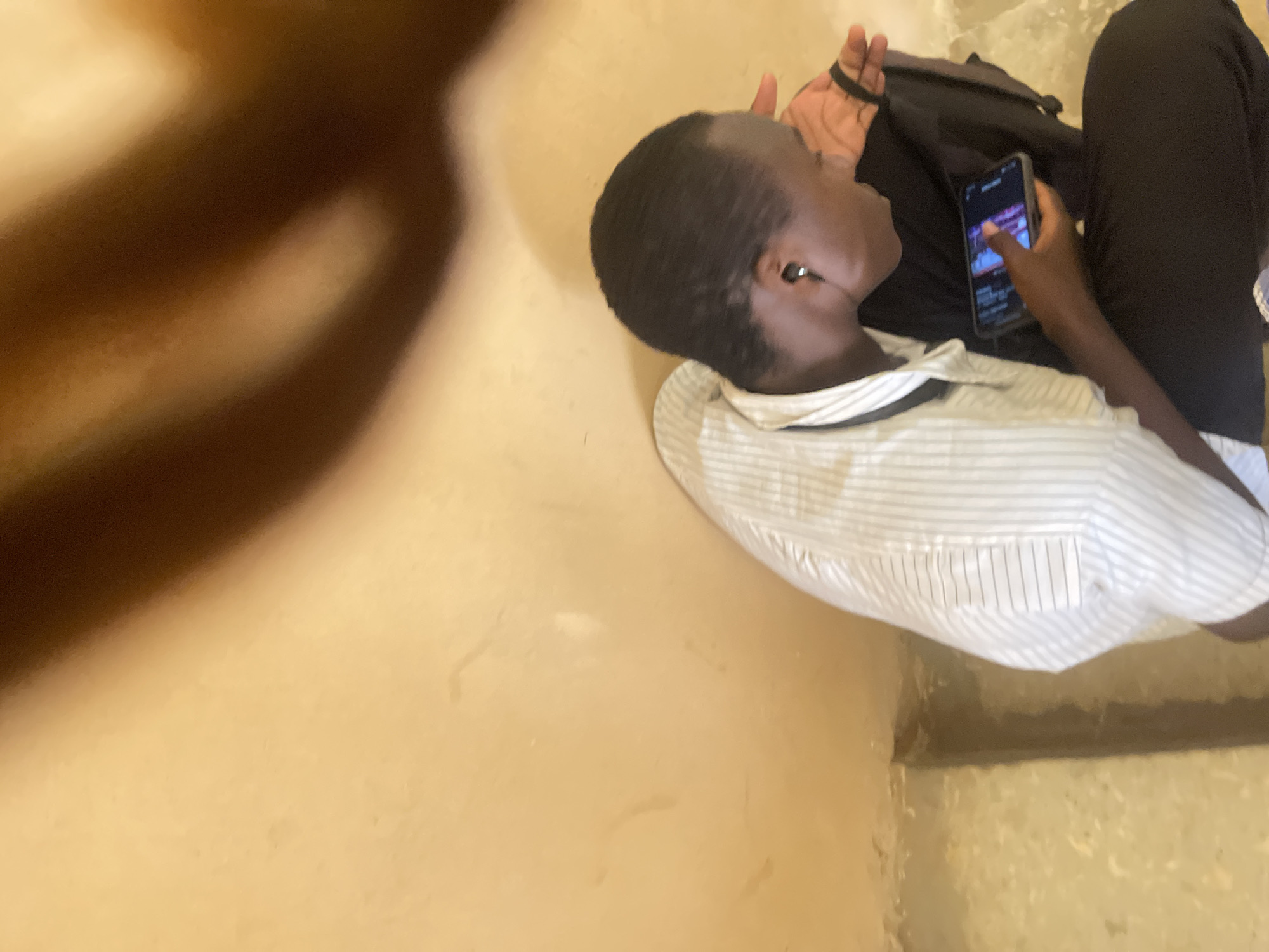 A person is shown from a high angle, sitting on steps with a beige wall behind them and a darker area to the left. The person is wearing a white pinstripe shirt and black pants. They have black earbuds in and are holding a smartphone in their right hand, looking down at it. A black backpack is visible behind them. The scene appears to be outdoors, possibly in a public space like a bus station or an outdoor waiting area. The overall lighting is warm and suggests daylight.