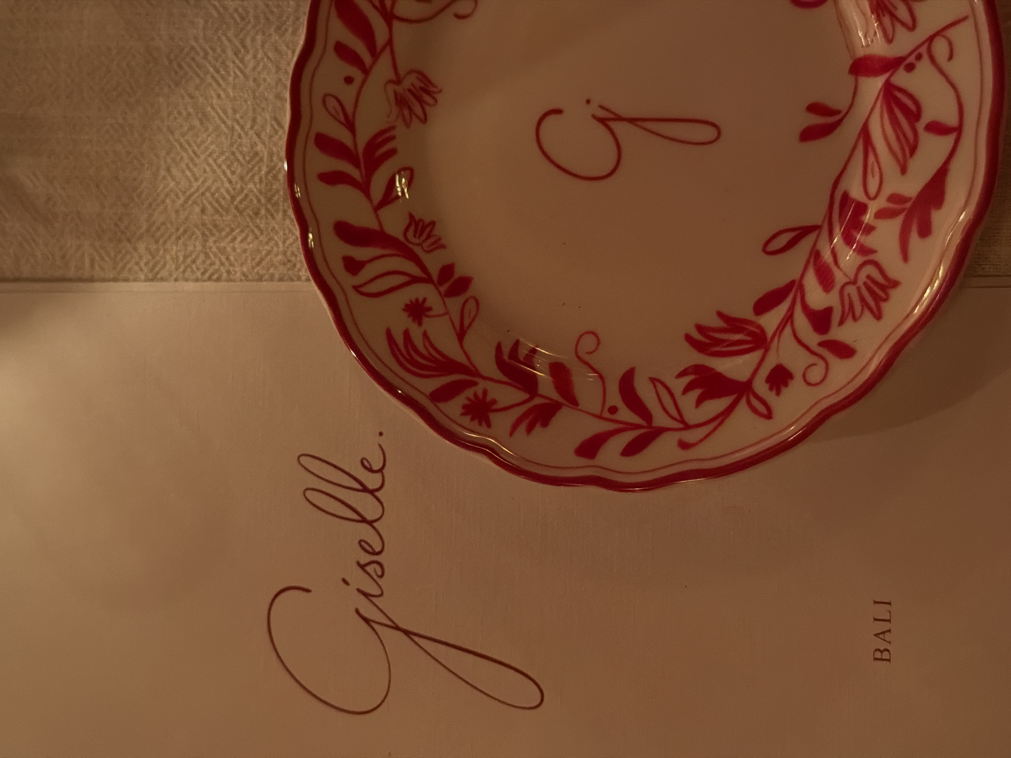 The image depicts a setting in Bali, specifically North Kuta, Indonesia, as indicated by the word "BALI" printed on a menu or card. The name "Giselle." is also printed on the menu/card in cursive script, likely referring to a person or establishment. A white plate with a red floral pattern and a red letter "J" in the center rests on the menu. Part of a textured white surface, possibly a tablecloth, is visible in the upper left corner. The scene is dimly lit, resulting in warm, subdued tones.