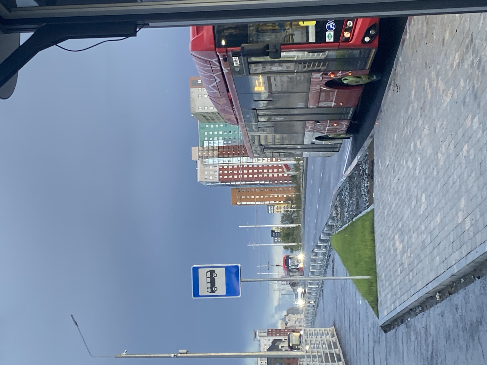 The image depicts a street scene in Perm, Russia, under a cloudy sky. A red bus is parked at a bus stop, visible on the right side of the frame. The bus stop sign, a blue square with a white bus silhouette, is prominently displayed in the center left. In the background, several multi-story residential buildings with colorful facades are visible, suggesting an urban setting. The road appears wet, indicating recent rain, and there is a paved sidewalk with a small patch of grass beside it. Other vehicles, including a tram and cars, can be seen on the road in the distance. The overall atmosphere is overcast and slightly gloomy.