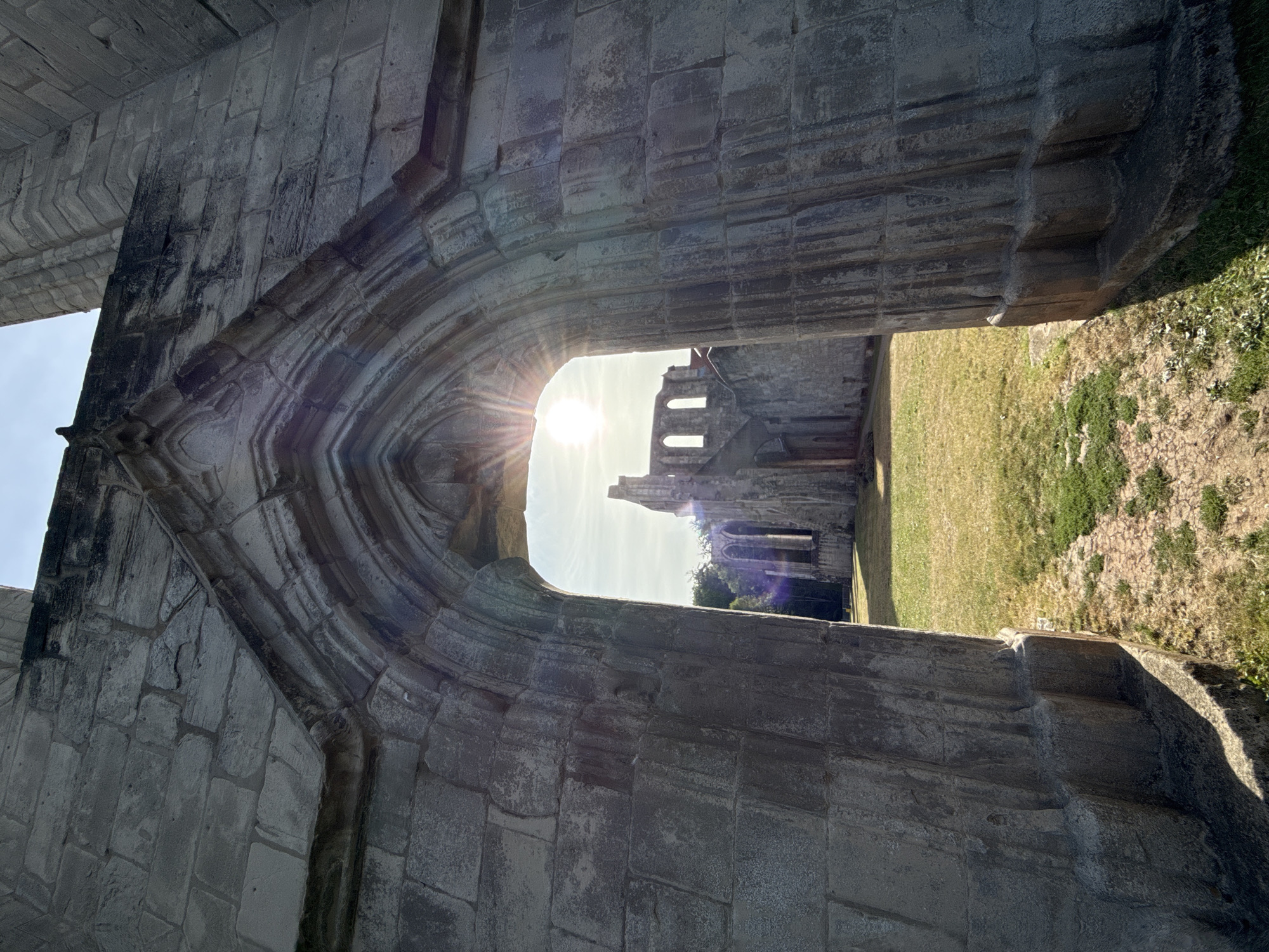 The image shows the ruins of Walkenried Abbey, located in Walkenried, Germany. The viewpoint is from inside a stone archway, looking out towards the remaining structure of the abbey. The sun is shining brightly through the archway, creating lens flare and highlighting the weathered stone walls of the ruins. The foreground features the rough texture of the stone arch, while the background shows a grassy area with more stone structures and the sky. No people are visible in the image.