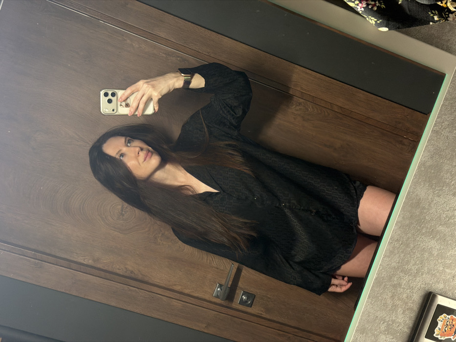 The image displays a mirror selfie of a woman, oriented diagonally across the frame with her head towards the upper left and feet towards the lower right.
The woman has long, straight, dark brown hair that cascades over her shoulders and across her chest. She is looking upwards and slightly to her right, with a neutral expression and light-colored eyes. She is wearing a dark (likely black) long-sleeved button-down shirt or blouse, which features a subtle, tonal patterned texture, possibly a very subdued animal print or geometric design. The garment has several small, dark buttons visible down the front. A watch with a wide, metallic (gold or bronze-toned) band is visible on her left wrist. Her right hand holds a light-colored smartphone with a transparent or light-toned case, positioned in front of her face as she takes the photo. Below the hem of her shirt, portions of her upper thighs are visible, suggesting the shirt might be worn as a dress or paired with very short bottoms.
The background, which is a reflection in the mirror, primarily consists of a wooden surface. This surface has a distinct grain pattern, including some circular "eyes" indicative of wood knots, suggesting it's a wooden door or wall paneling. A modern, metallic lever-style door handle is visible on this wooden surface. Above the wooden section, there's a dark horizontal strip, and further up, a lighter, possibly gray, wall or ceiling.
In the top right corner of the overall image, a small section of a floral-patterned fabric (dark background with light-colored flowers) is partially visible, likely a piece of clothing or fabric outside the mirror's main reflection. In the bottom right corner, on what appears to be a gray countertop or shelf adjacent to the mirror, a small rectangular object is visible with several stickers applied to its surface. One sticker clearly depicts an orange and brown cartoon-like animal character.