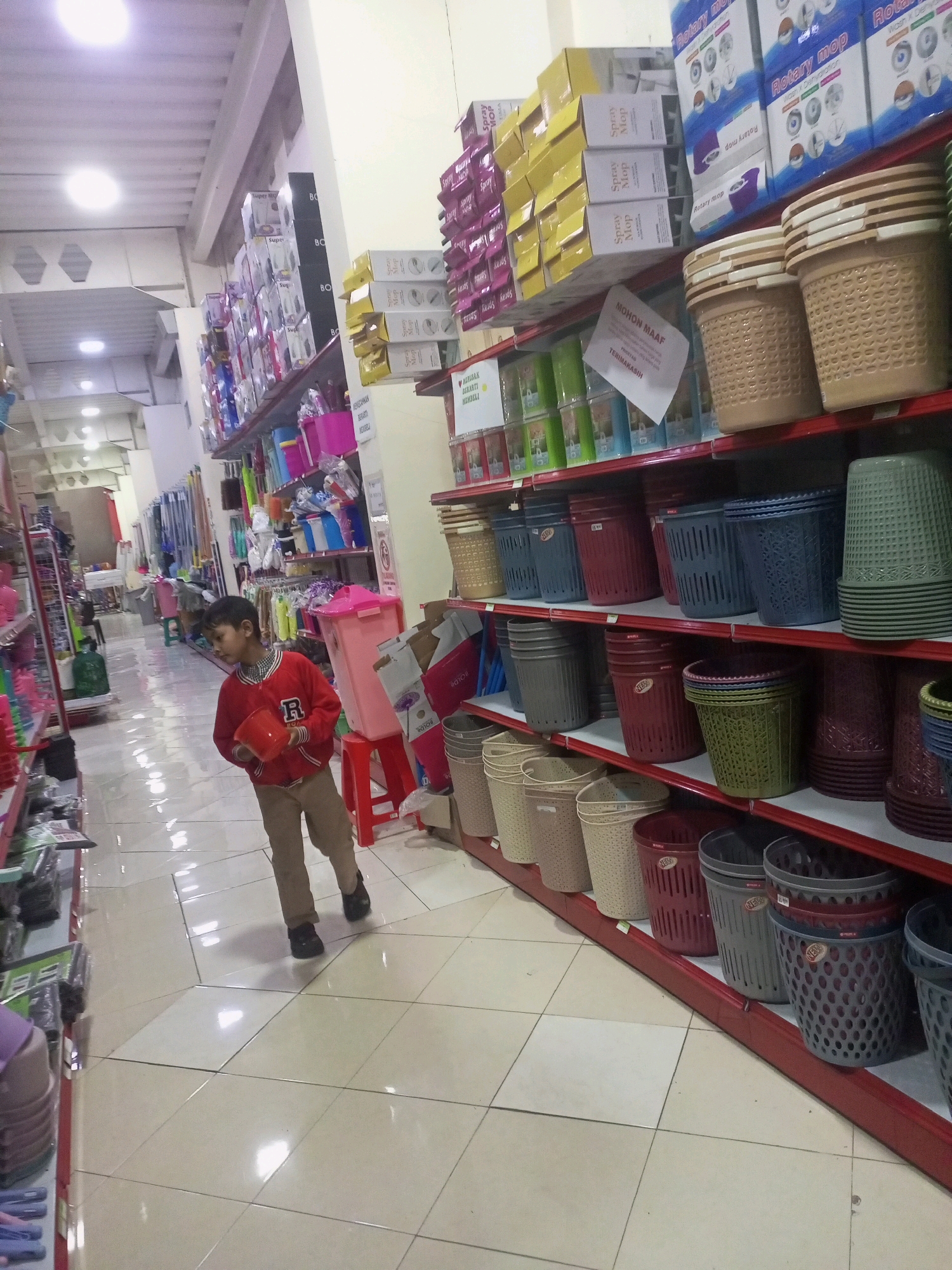 The image depicts the interior of a retail store, likely a hardware or household goods store, in Ngasem, Indonesia. The store is well-stocked with various plastic household items, predominantly baskets and storage containers, displayed on red metal shelving units. In the foreground, a young boy wearing a red jacket and beige pants is walking, holding a red plastic bucket. The store's flooring is tiled, reflecting the overhead fluorescent lighting. The shelves are organized with multiple rows of colorful plastic baskets, varying in size, shape, and color, including beige, blue, dark red, green, and gray. Some shelves also display packaged goods, including what appear to be cleaning supplies and mops, with labels in Indonesian. A sign with "MOHON MAAF" (meaning "Excuse me" or "Sorry") and "TERIMAKASIH" (meaning "Thank you") is visible on one of the shelves, suggesting it's a general announcement or apology to customers. The overall environment suggests a typical Indonesian market or discount store setting.