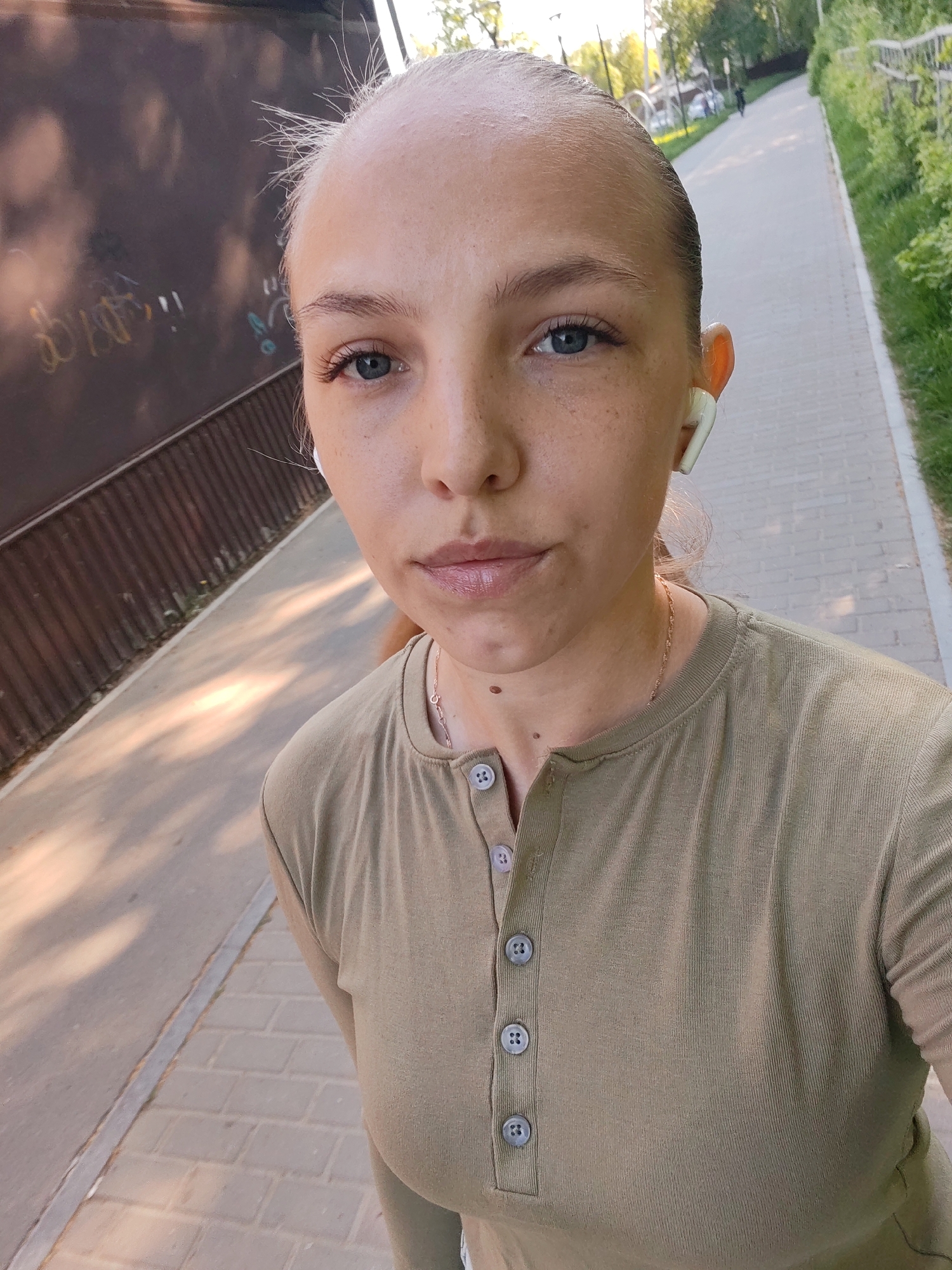 A fair-skinned woman with blue eyes and freckles is taking a selfie while walking on a paved pathway. She is wearing a buttoned, olive-green shirt and white wireless earbuds. Behind her is a dark brown fence with graffiti and foliage. The pathway continues into the distance, with trees and greenery visible along the side. The setting appears to be an outdoor public space in Chaikovsky, Russia.