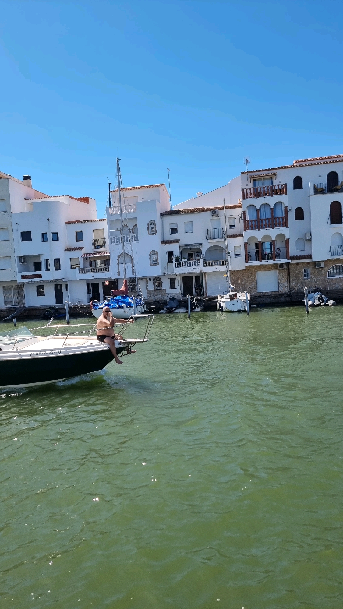 In Castelló d'Empúries, Spain, a man sits on the bow of a moving motorboat, facing left. He is wearing dark swim shorts and appears to be bald. The boat is a dark green and white vessel with a license plate that reads "BA-2-55-10". The boat is in a canal or waterway, with a row of white buildings lining the right side of the frame. These buildings have multiple stories, balconies with railings, and arched windows. Several other boats, including sailboats and smaller dinghies, are docked or moored in the background. The water is a murky green, and the sky is clear blue.
