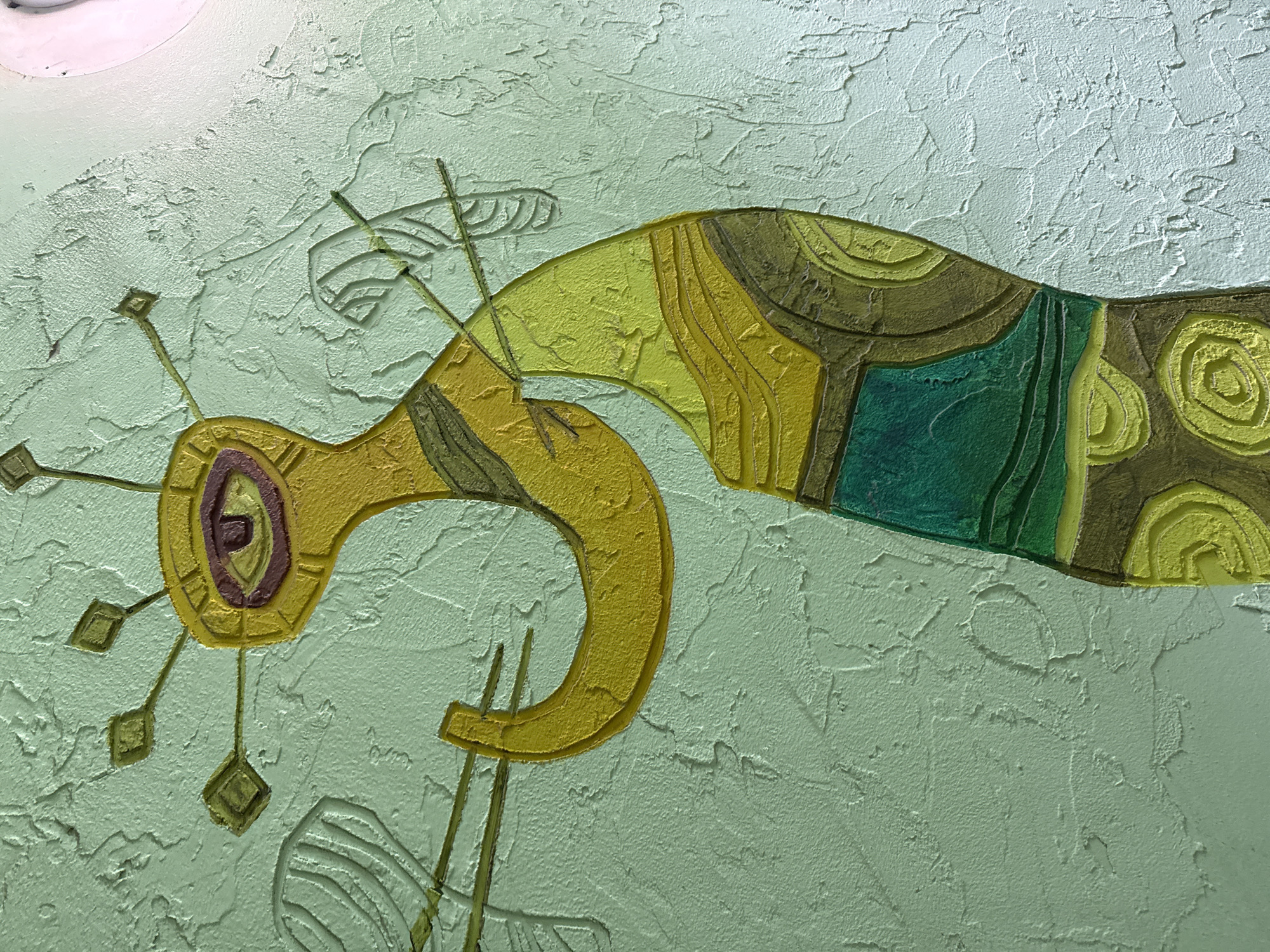 A textured, pale green wall showcases an abstract, stylized artwork. The piece is dominated by a yellow, elongated shape reminiscent of a bird or dragon, with darker green and brown details suggesting patterns and divisions. The head of the creature is a ring-shaped form with linear "antennae" ending in square shapes. Behind it are faint sketched lines suggesting wings or other appendages. The background is a textured surface, possibly stucco or a similar material. The artwork appears to be integrated into the wall's surface. The location is unknown and can not be inferred from this image.