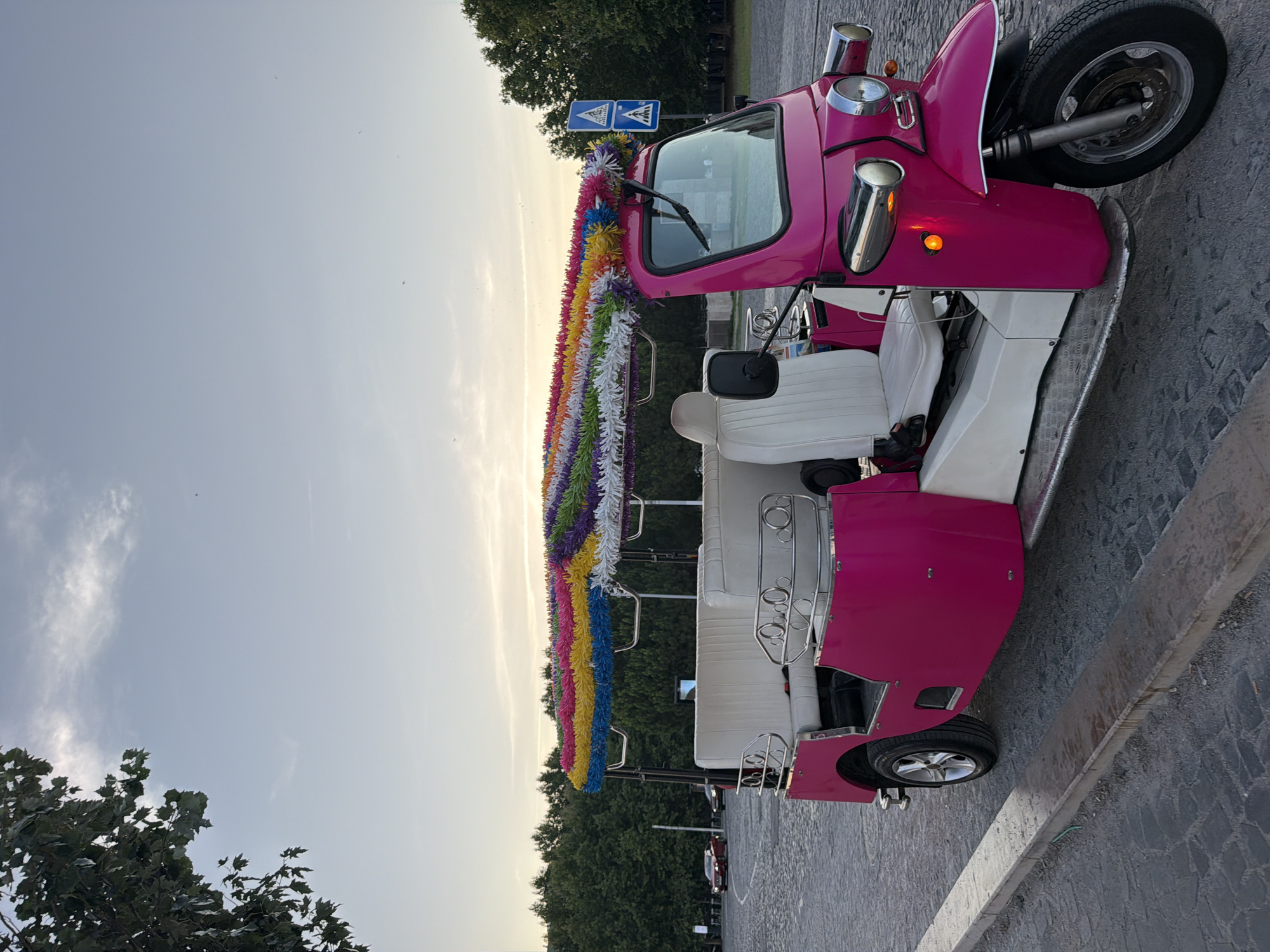 A pink and white three-wheeled vehicle, resembling a tuk-tuk, is parked on a cobblestone street in Lisbon, Portugal. The vehicle is decorated with colorful streamers along its roof. White seats are visible inside the passenger area. To the right, a traffic sign with pedestrian symbols is posted. In the background, a line of trees forms a dense backdrop against a pale sky with wispy clouds. A red car is visible further down the street to the left. The overall scene suggests an outdoor setting, possibly a tourist area, during the day.