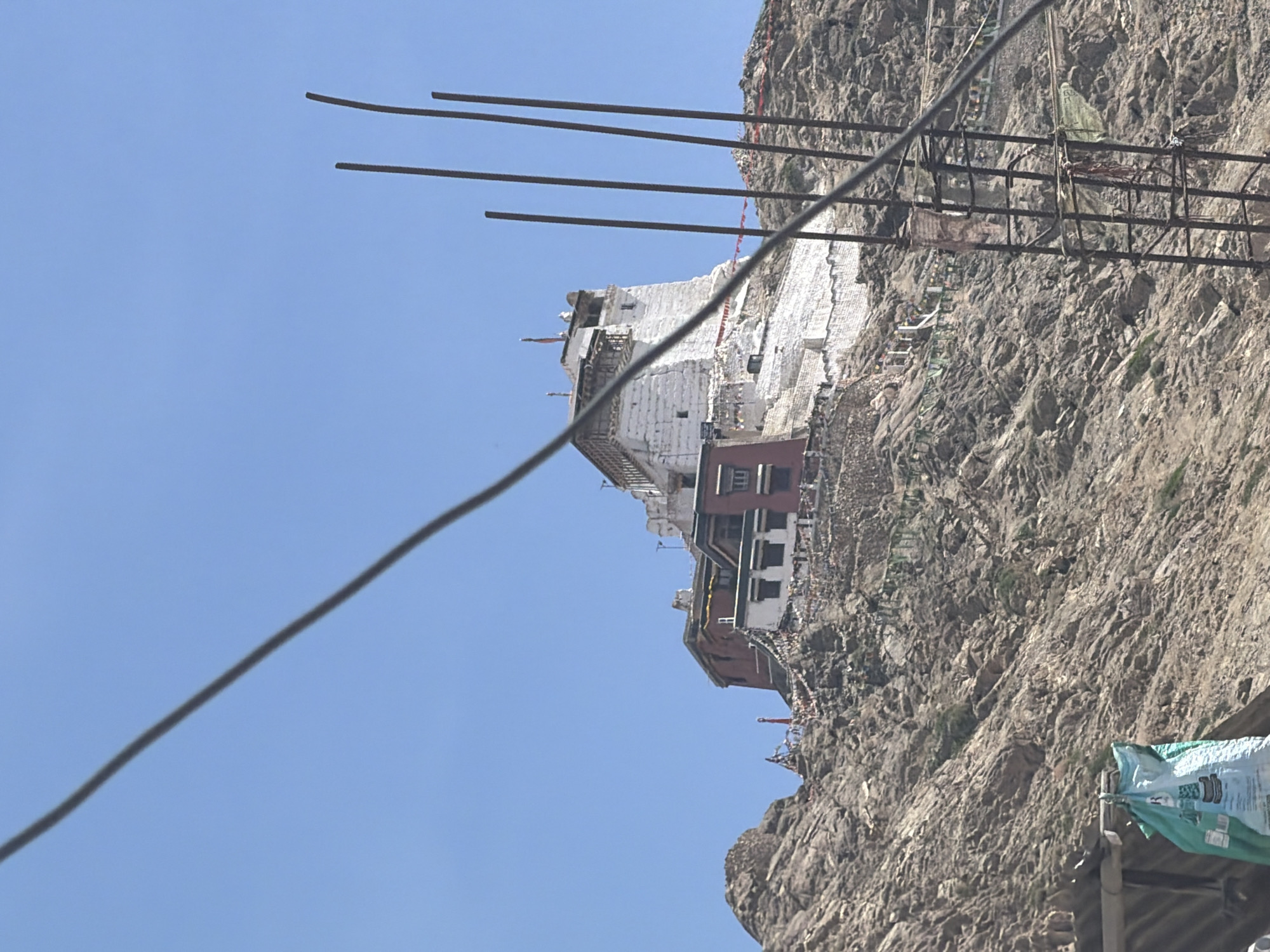 The image depicts the Leh Palace, a historic monument located in the city of Leh, Ladakh, India. The palace is perched atop a rocky hill, its white and red structures contrasting with the clear blue sky. Prayer flags, adorned with colorful inscriptions, are strung across the hillside, adding a touch of vibrancy to the rugged landscape. In the foreground, construction materials, including rebar and a blue tarp, suggest ongoing development or maintenance work near the base of the hill. The overall scene captures the grandeur of the palace against a backdrop of natural beauty and human activity.