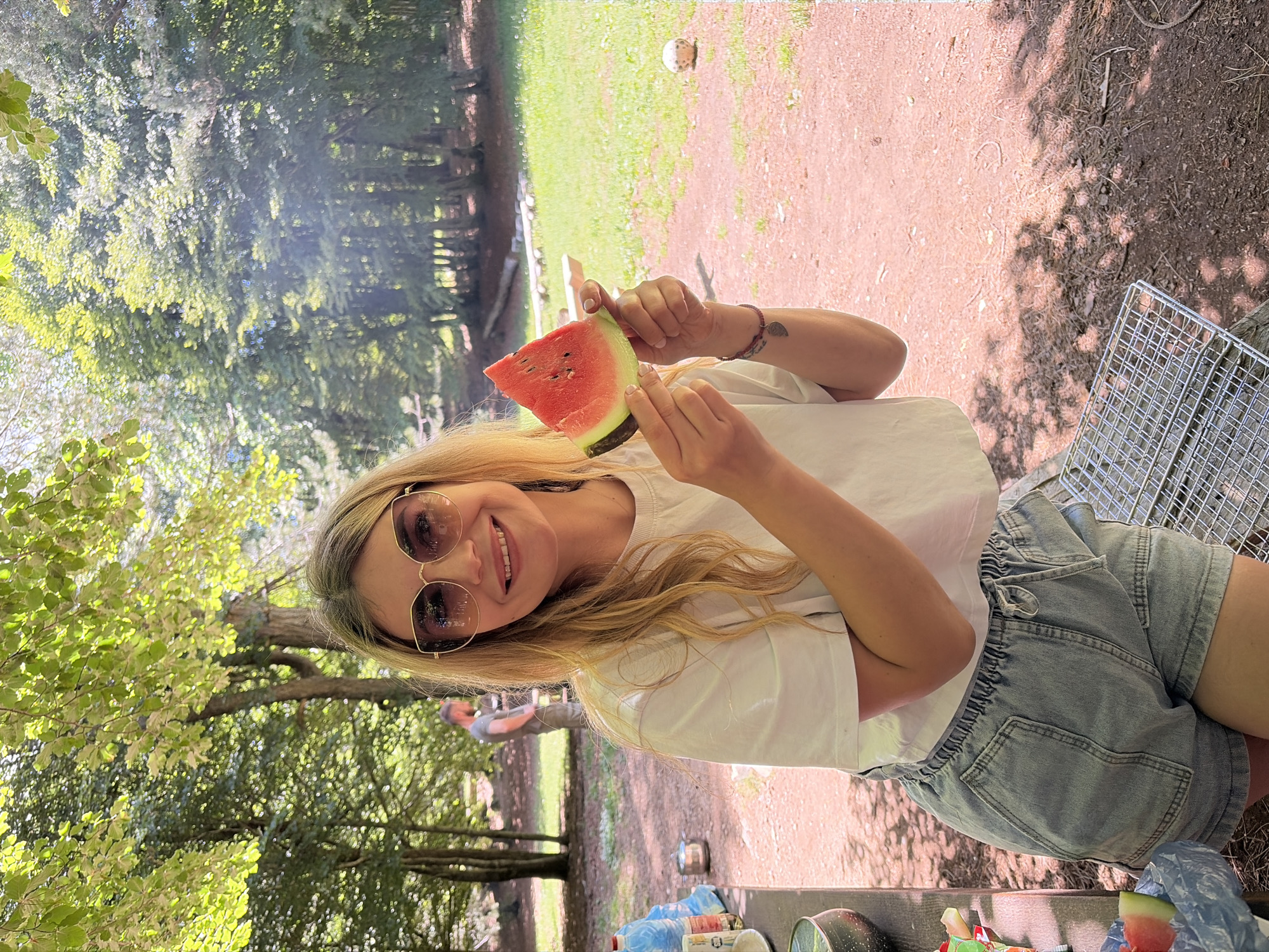 The image depicts an outdoor gathering in a wooded area within Dolovi, Montenegro, under bright daylight. A blonde female with sunglasses and a white t-shirt stands centrally, smiling at the viewer while holding a triangular slice of red watermelon with black seeds. She wears light blue denim shorts and a dark bracelet on her left wrist. To her left, partially visible on a picnic table, are two large blue water bottles, a small silver pot, and a dark speckled mug. A blue plastic bag containing another watermelon slice rests near the table. In the mid-ground on the left, a male figure wearing a grey t-shirt and dark trousers is visible from the back, facing away from the camera. The ground in the foreground and mid-ground consists of reddish-brown dirt with scattered dry leaves and twigs. A metal barbecue grill rack lies on the ground in the bottom right foreground. Beyond the dirt path, a bright green grassy area extends into the background, where a deflated or dirty white soccer ball is visible on the grass and several rough logs or benches are arranged. The scene is heavily treed, with dense green foliage creating dappled sunlight and shadows on the ground.